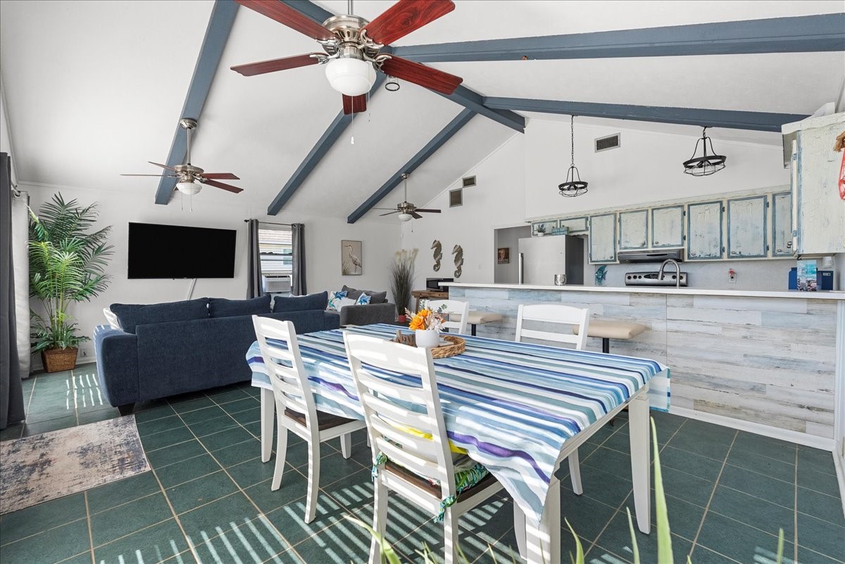 324 Shark Lane Surfside Beach, TX 77541 - Photo 15 of 49 This open-concept living space features high vaulted ceilings with exposed beams, ceiling fans, and natural light. The area includes a cozy living room with a large TV, a dining area with a striped tablecloth, and a kitchen with rustic cabinets and a breakfast bar, all set on dark tiled flooring.