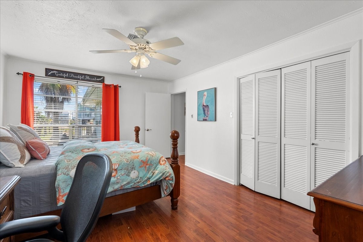 324 Shark Lane Surfside Beach, TX 77541 - Photo 22 of 49 This bright bedroom features hardwood floors, a ceiling fan, and ample natural light from a large window with red curtains. It includes a cozy bed, built-in closet, and a small desk, making it ideal for relaxation and productivity.