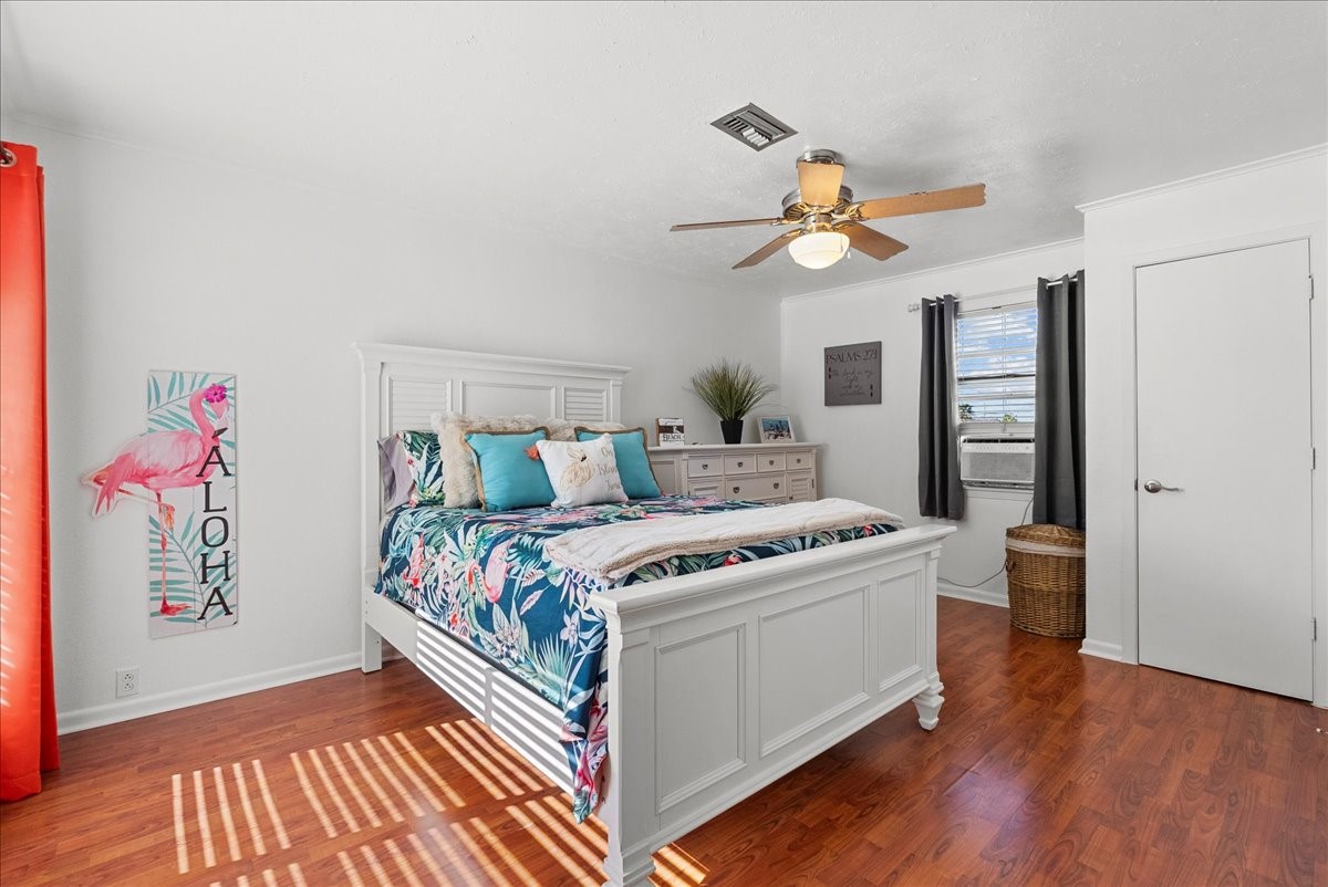 324 Shark Lane Surfside Beach, TX 77541 - Photo 23 of 49 This bright and airy bedroom features hardwood look floors, a ceiling fan, and a large window with natural light. The room is tastefully decorated with a tropical theme, including a colorful bedspread and wall art. A white bed frame and dresser provide ample storage and complement the room's fresh, clean aesthetic.