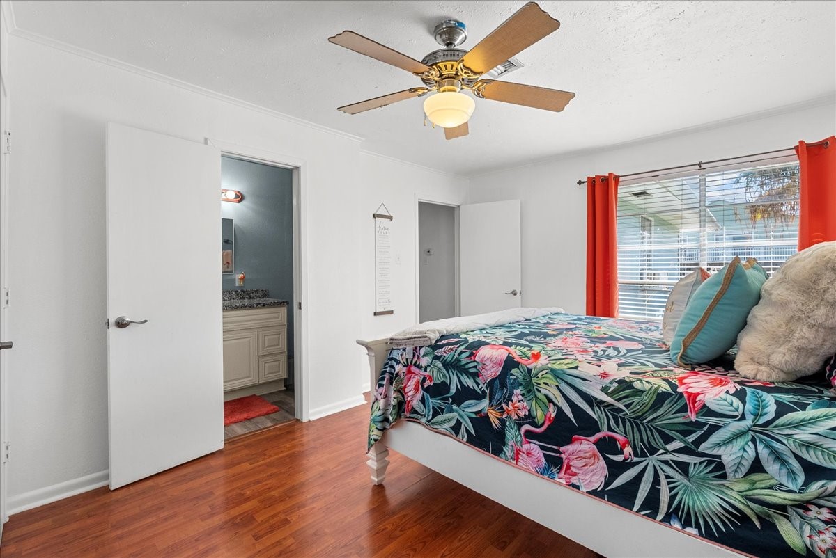324 Shark Lane Surfside Beach, TX 77541 - Photo 24 of 49 This bright bedroom features hardwood look flooring, a ceiling fan, and ample natural light through a large window with vibrant curtains. It has an attached bathroom with a modern vanity, offering convenience and style.