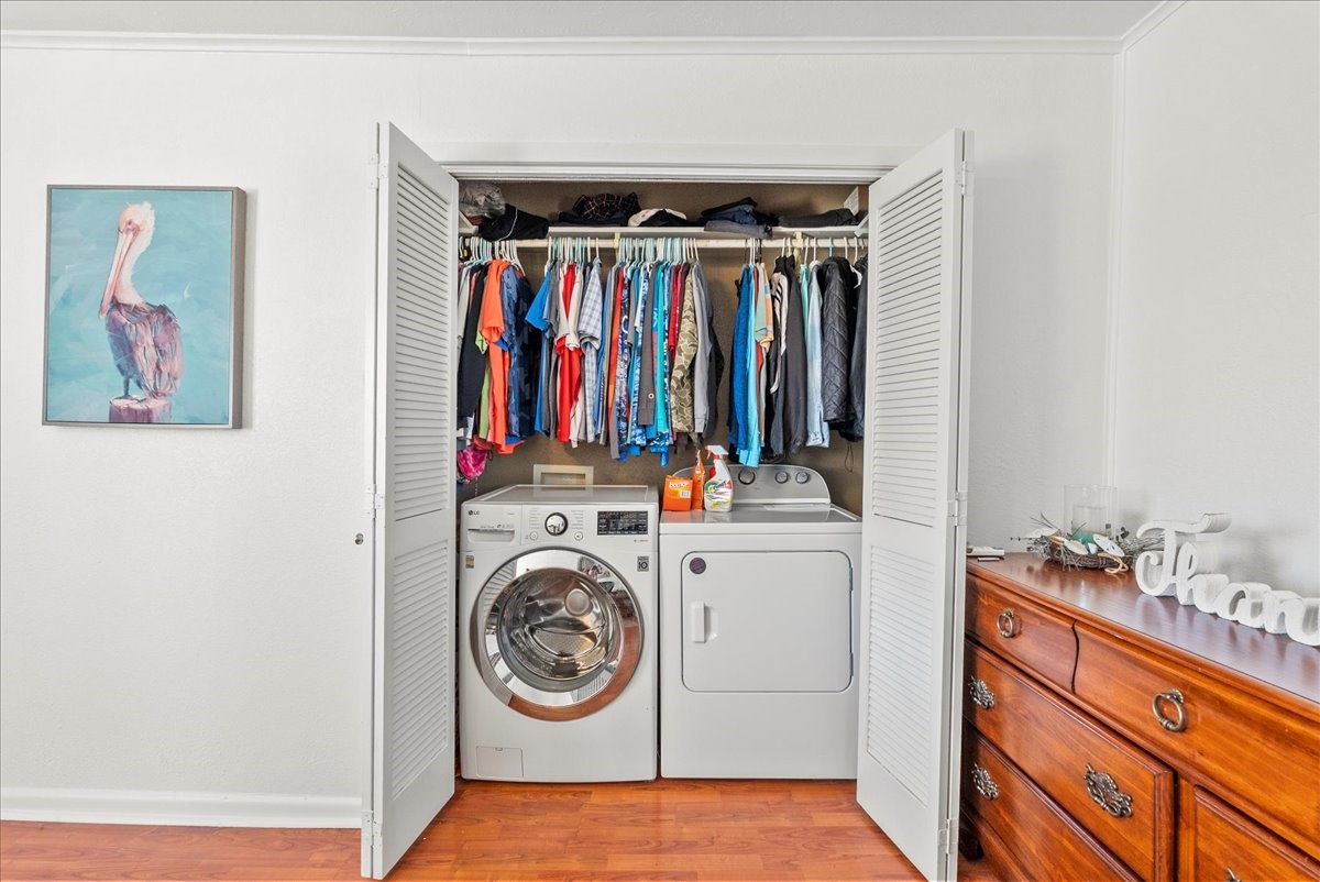 324 Shark Lane Surfside Beach, TX 77541 - Photo 29 of 49 This photo shows a compact laundry area with a washer and dryer neatly tucked into a closet with open louvered doors. Above, there's a rod with hanging clothes. The room features hardwood look flooring, a wooden dresser, and a wall painting, offering a practical and organized space.
