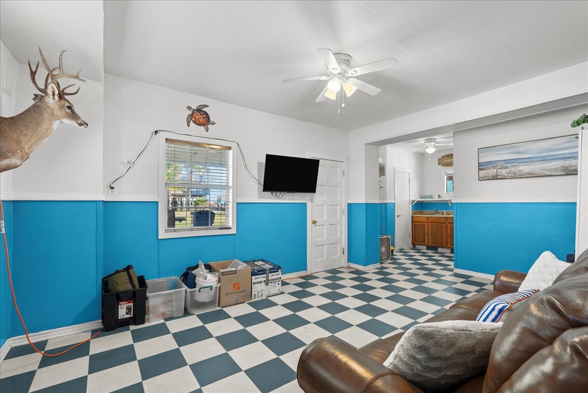 324 Shark Lane Surfside Beach, TX 77541 - Photo 35 of 49 This room features a bright, checkerboard floor and vibrant blue and white walls. It includes ceiling fans, a mounted TV, and a window for natural light. Decor elements include wall-mounted art and a deer head. The space leads into an area with visible cabinet storage.
