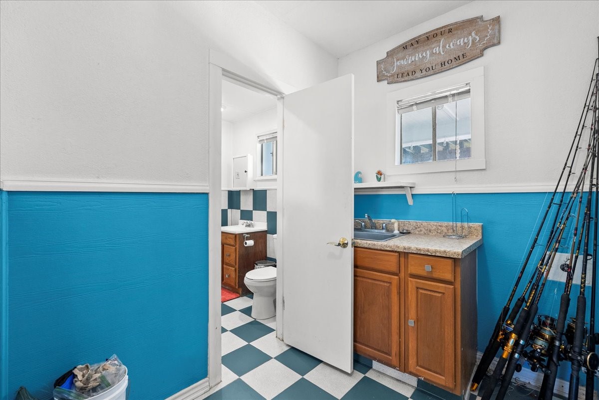 324 Shark Lane Surfside Beach, TX 77541 - Photo 36 of 49 This photo shows a compact laundry room or utility area with bright blue and white walls. There is a small countertop with a sink and wooden cabinets. The flooring features a checkered pattern.