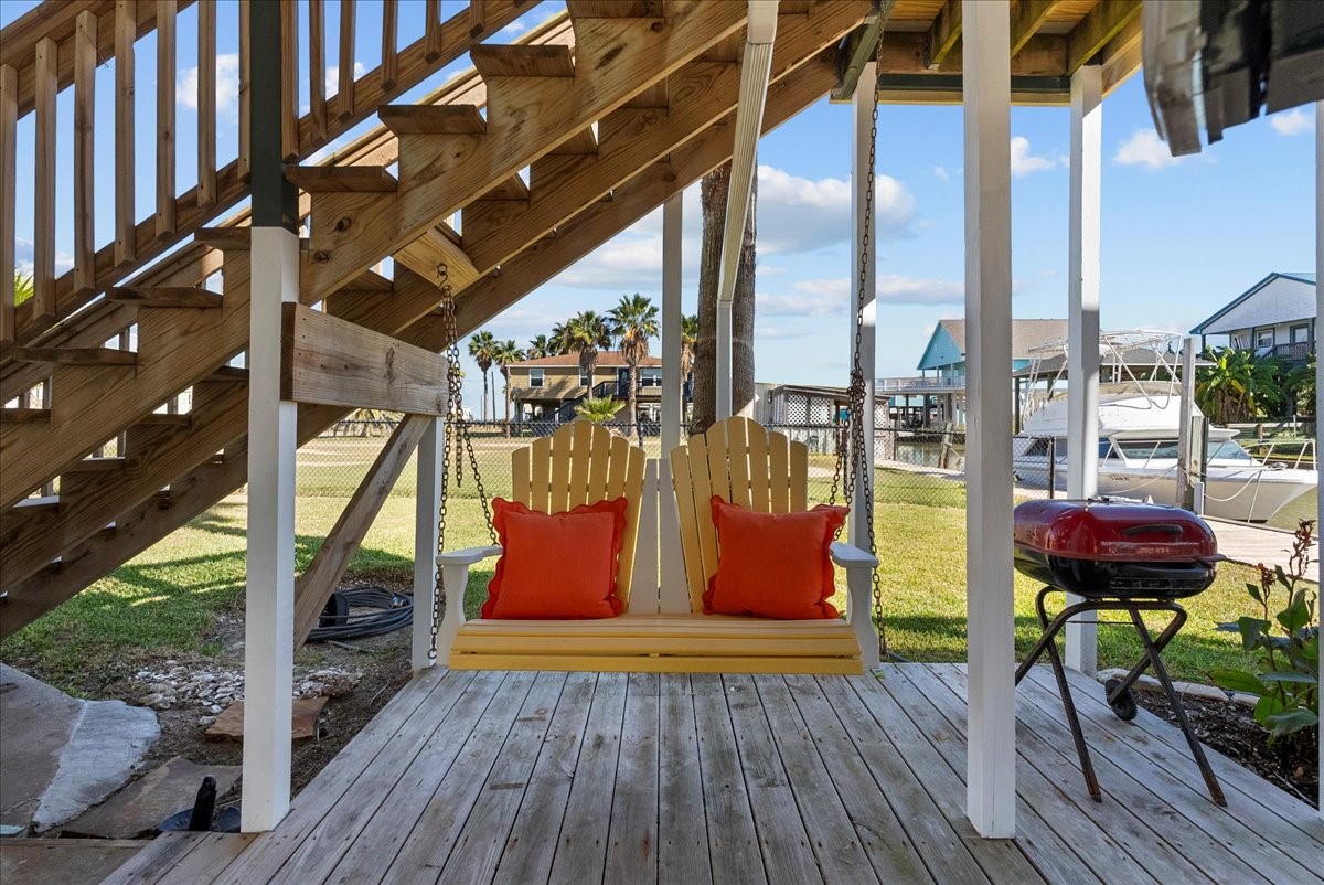 324 Shark Lane Surfside Beach, TX 77541 - Photo 41 of 49 This inviting outdoor space features a cozy wooden swing with bright orange cushions, set under a staircase. It overlooks a tranquil waterfront, perfect for relaxation and entertaining, complete with a grill nearby.