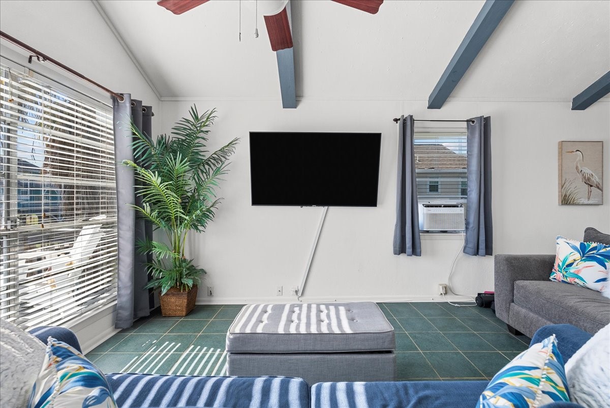 324 Shark Lane Surfside Beach, TX 77541 - Photo 8 of 49 Bright living room with large windows and natural light. Features include a mounted TV, comfortable gray sofas with colorful cushions, and a ceiling fan. A potted plant adds a touch of greenery.