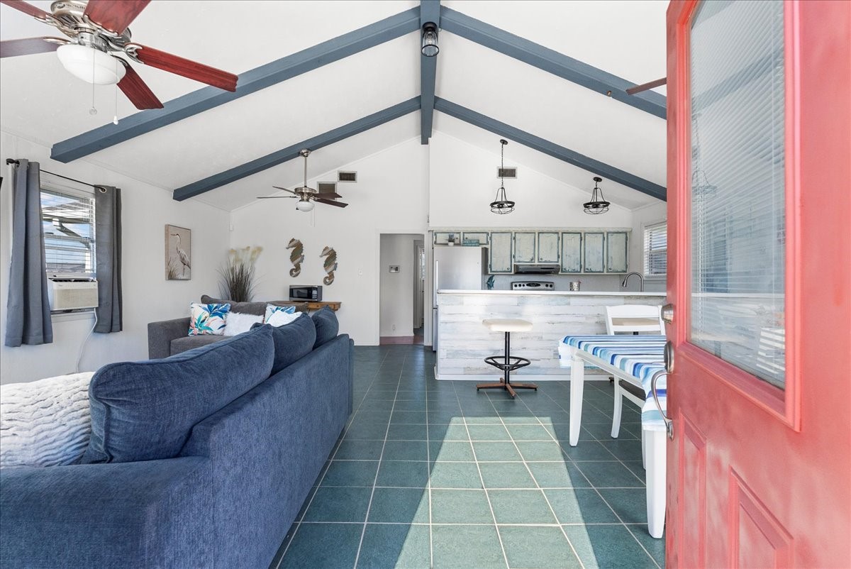 324 Shark Lane Surfside Beach, TX 77541 - Photo 9 of 49 This inviting home features an open-concept living area with vaulted ceilings and exposed beams. It includes a cozy navy sofa, modern ceiling fans, and large windows for natural light. The kitchen has rustic cabinets and a breakfast bar, creating a welcoming space for entertaining.