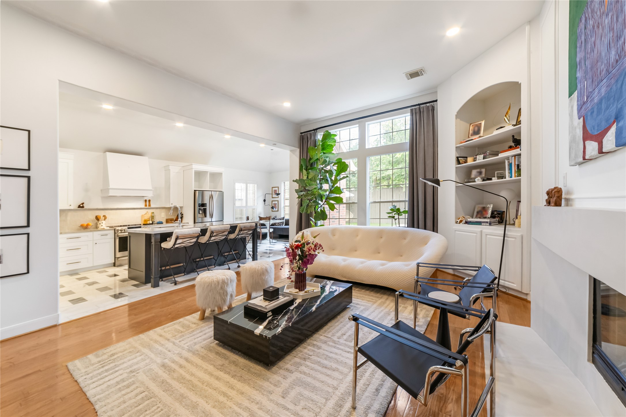 This photo showcases a bright, open-concept living space with hardwood floors, featuring a modern kitchen with a large island and stainless steel appliances. The living area offers a inviting area with a fireplace and built ins. Large windows provide ample natural light and a view of the backyard.