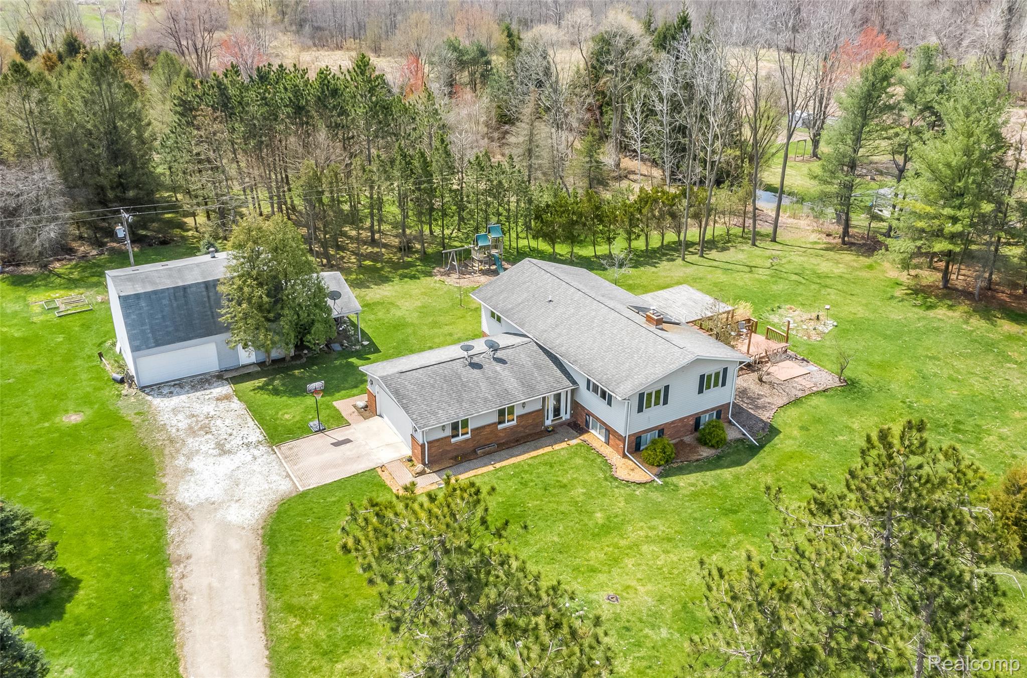 15395 Osius Road Chelsea, MI 48118 - Photo 45 of 55 Expansive property featuring a residence with a light gray roof and brick siding, a detached garage, a gravel driveway, and mature trees surrounding the perimeter
