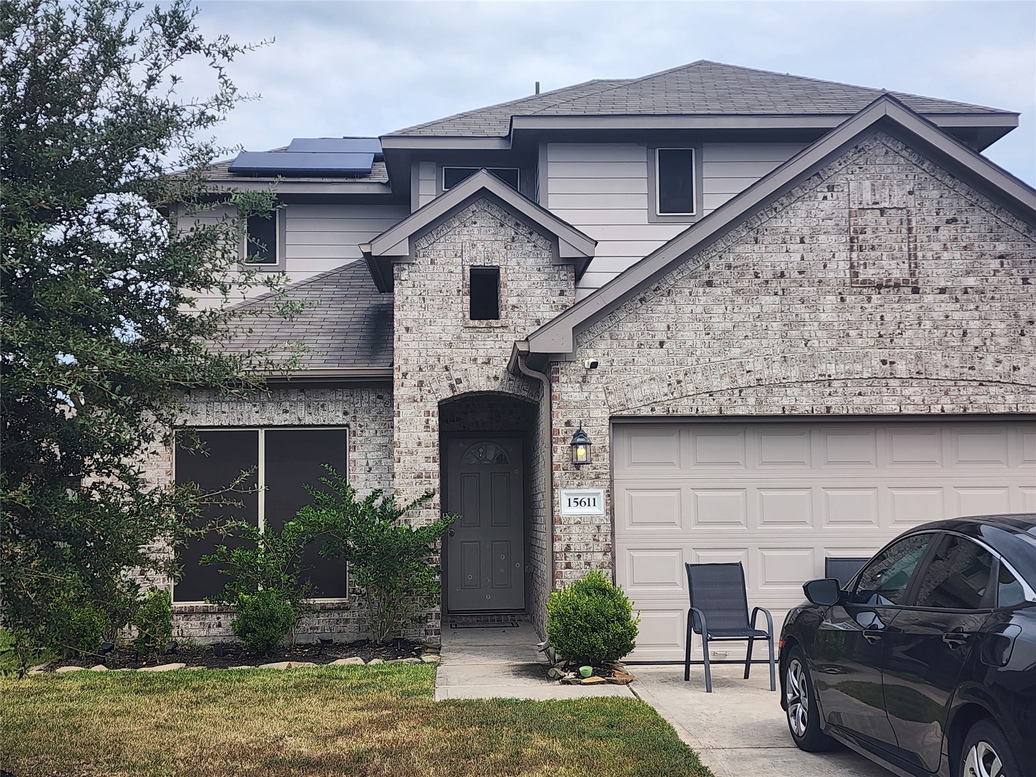 Two-story home features a brick exterior with a gabled roof and solar panels. It includes a two-car garage. The entrance is adorned with a single light fixture and a covered entryway.