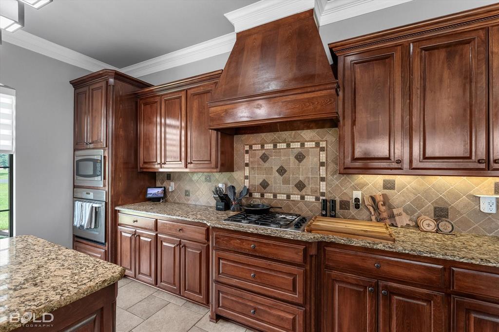 2220 Forest Hills Boulevard Haughton, LA 71037 - Photo 15 of 40 Kitchen featuring custom wood cabinetry, granite countertops, tile backsplash with decorative insert, built-in stainless steel appliances, and a gas cooktop with a custom wood range hood