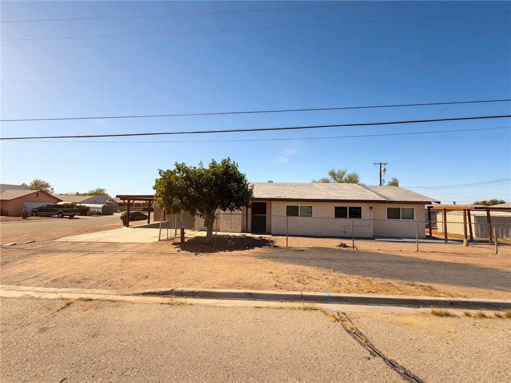 Ranch-style residence featuring a light-colored exterior, an attached carport, a chain-link fence, and a mature tree in the front yard