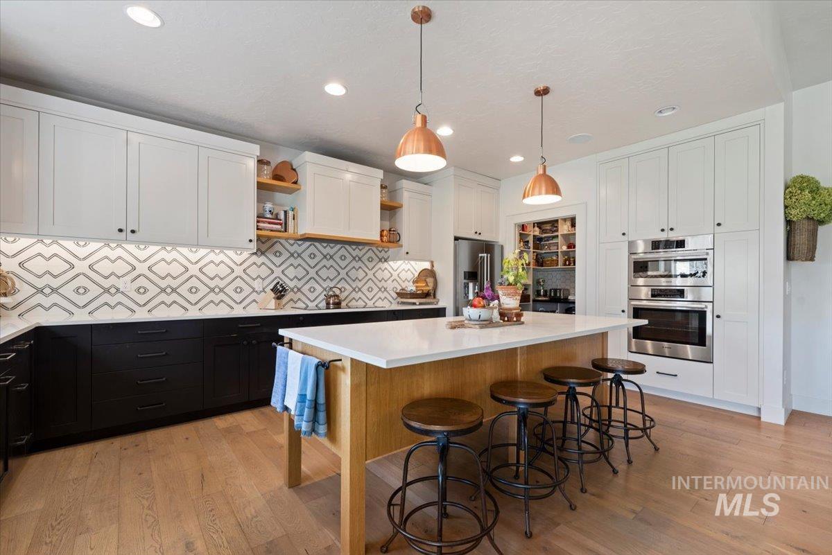 15164 Pinehurst Way Caldwell, ID 83607 - Photo 15 of 50 Gourmet kitchen featuring a Copper Sink, wood-finish island with a light-toned countertop, coordinated pendant lighting, two-tone cabinetry with matte black hardware, integrated stainless steel double ovens, and a patterned tile backsplash
