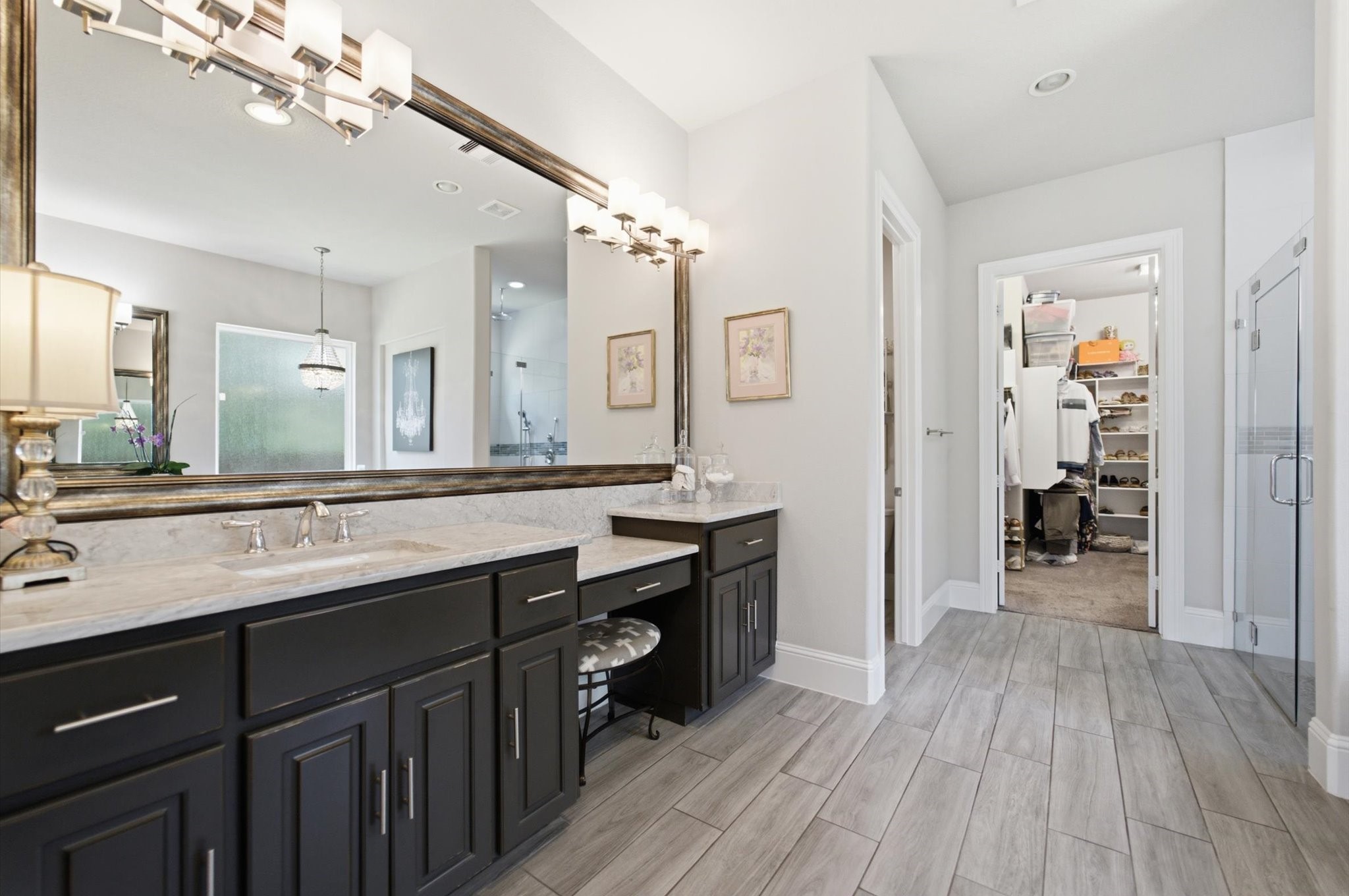 23722 Thortons Park Lane Spring, TX 77389 - Photo 13 of 48 This photo showcases a modern bathroom with a spacious double vanity featuring dark cabinetry and large mirrors. There's elegant lighting, a walk-in shower, and access to a roomy walk-in closet. Neutral tones and wood-like tile flooring provide a sleek, contemporary feel.