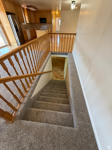 328 Heather Ridge Circle Sandy, UT 84070 - Photo 22 of 32 Carpeted staircase with wood railing, offering a glimpse of the kitchen featuring wood-finish cabinetry, stainless steel appliances, and a central island