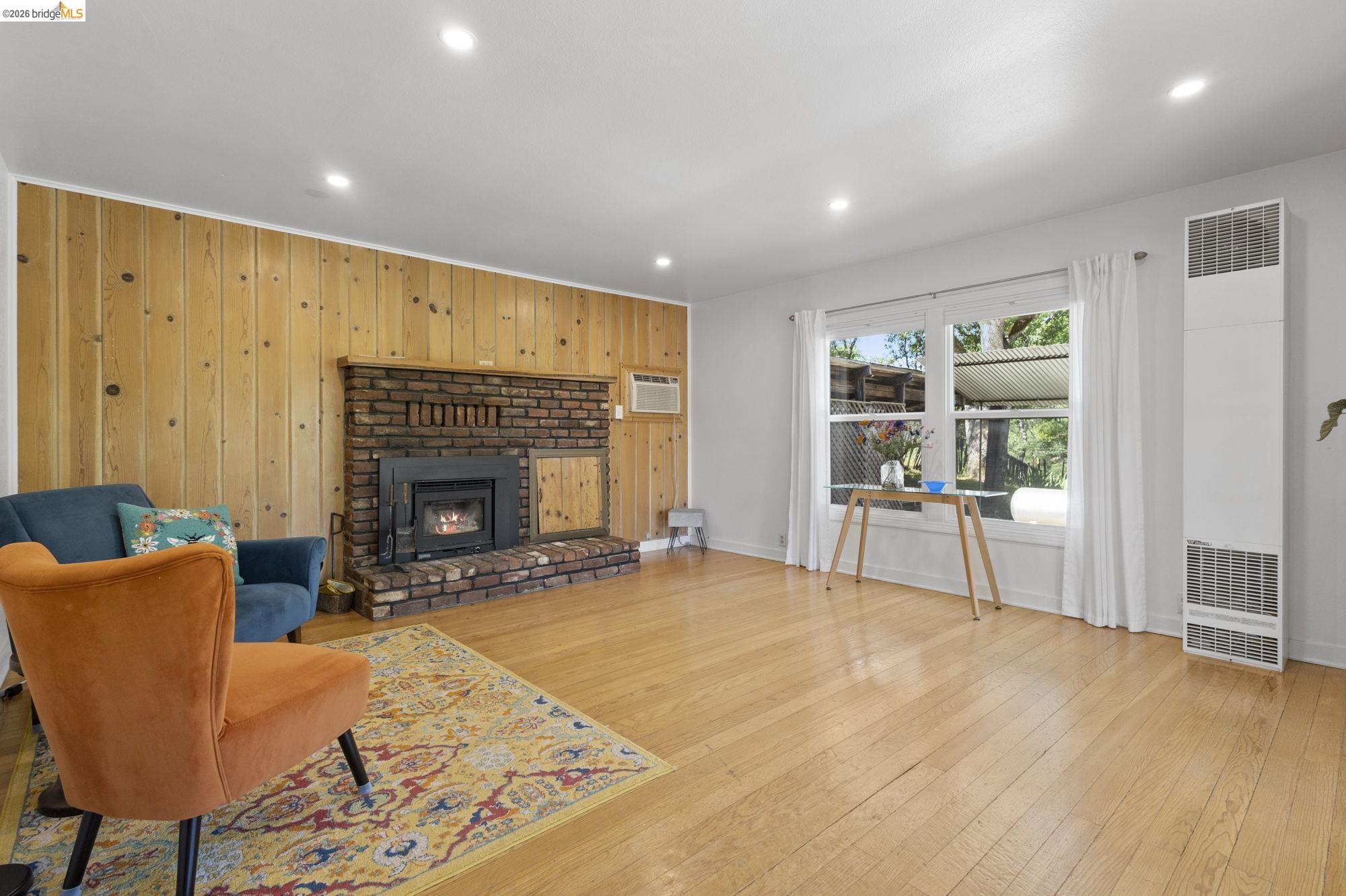18363 Yosemite Road Tuolumne, CA 95379 - Photo 7 of 34 Living area featuring wood-paneled accent wall, brick fireplace with insert, light wood-finish flooring, recessed ceiling lighting, and a large window with white curtain panels