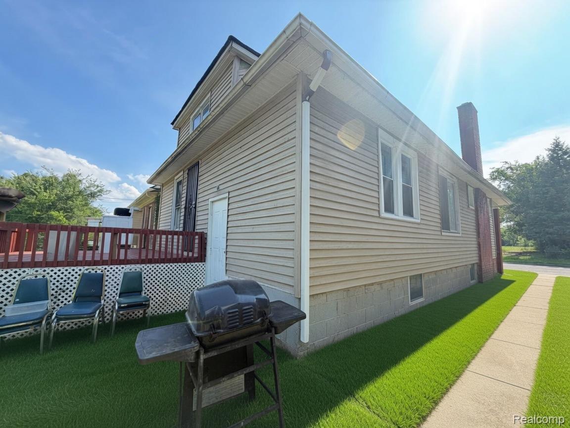 9940 Manor Detroit, MI 48204 - Photo 2 of 16 Exterior showcasing tan siding, a red deck with white lattice skirting, a concrete sidewalk, a brick chimney, and a green lawn