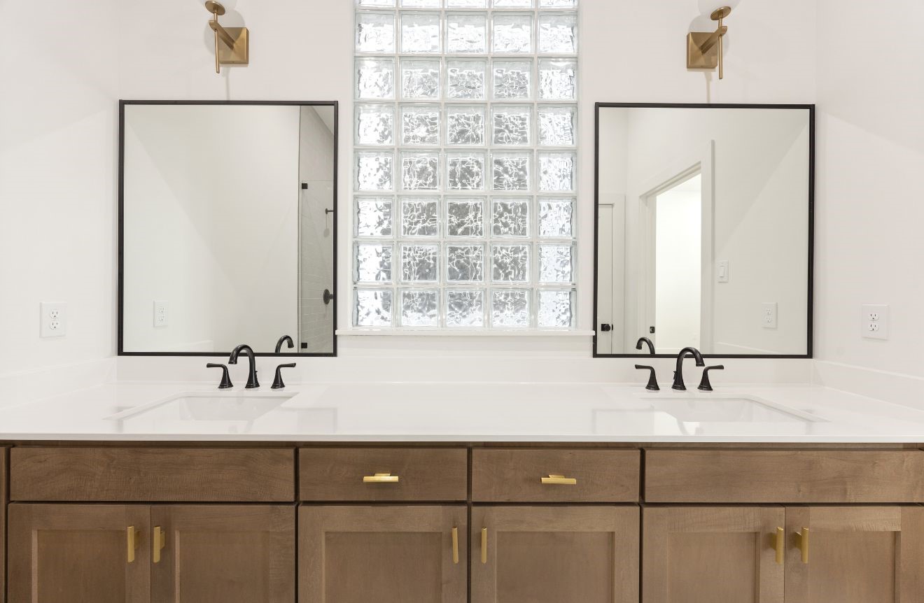 2121 Hoskins Drive, Unit C Houston, TX 77080 - Photo 27 of 34 Double vanity detail showcasing warm wood cabinetry, sleek white countertop, black framed mirrors, black faucets, brass-toned sconces, and a glass block window that brings in filtered natural light. The balanced design blends modern contrast with soft warmth for a stylish, spa-like bathroom setting.