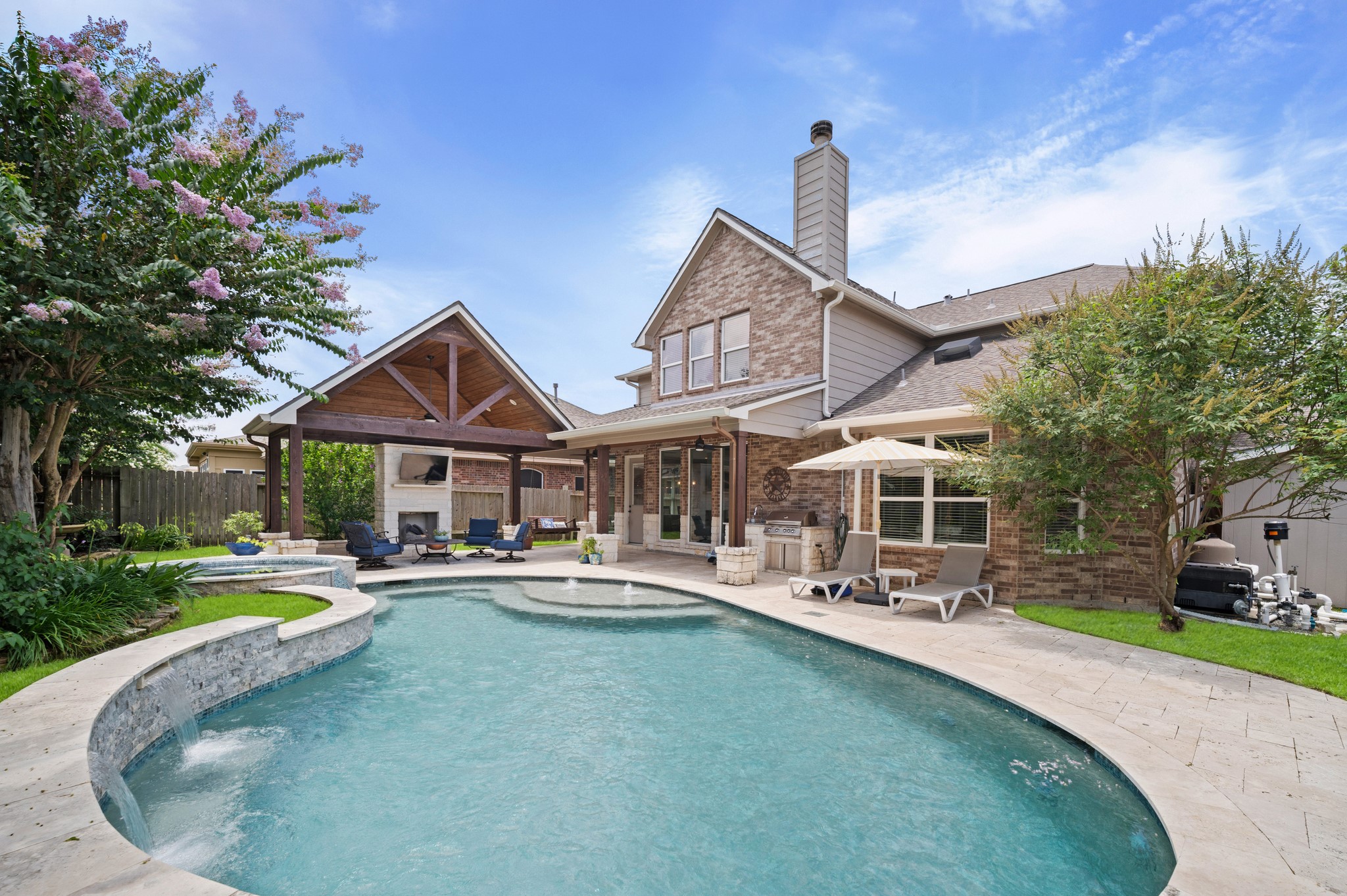 9507 Empress Crossing Drive Spring, TX 77379 - Photo 2 of 50 This photo showcases a luxurious backyard with a spacious swimming pool, a stone patio, and a covered outdoor living area complete with a fireplace and seating. The brick house features large windows and a well-maintained garden, creating an inviting and private retreat.