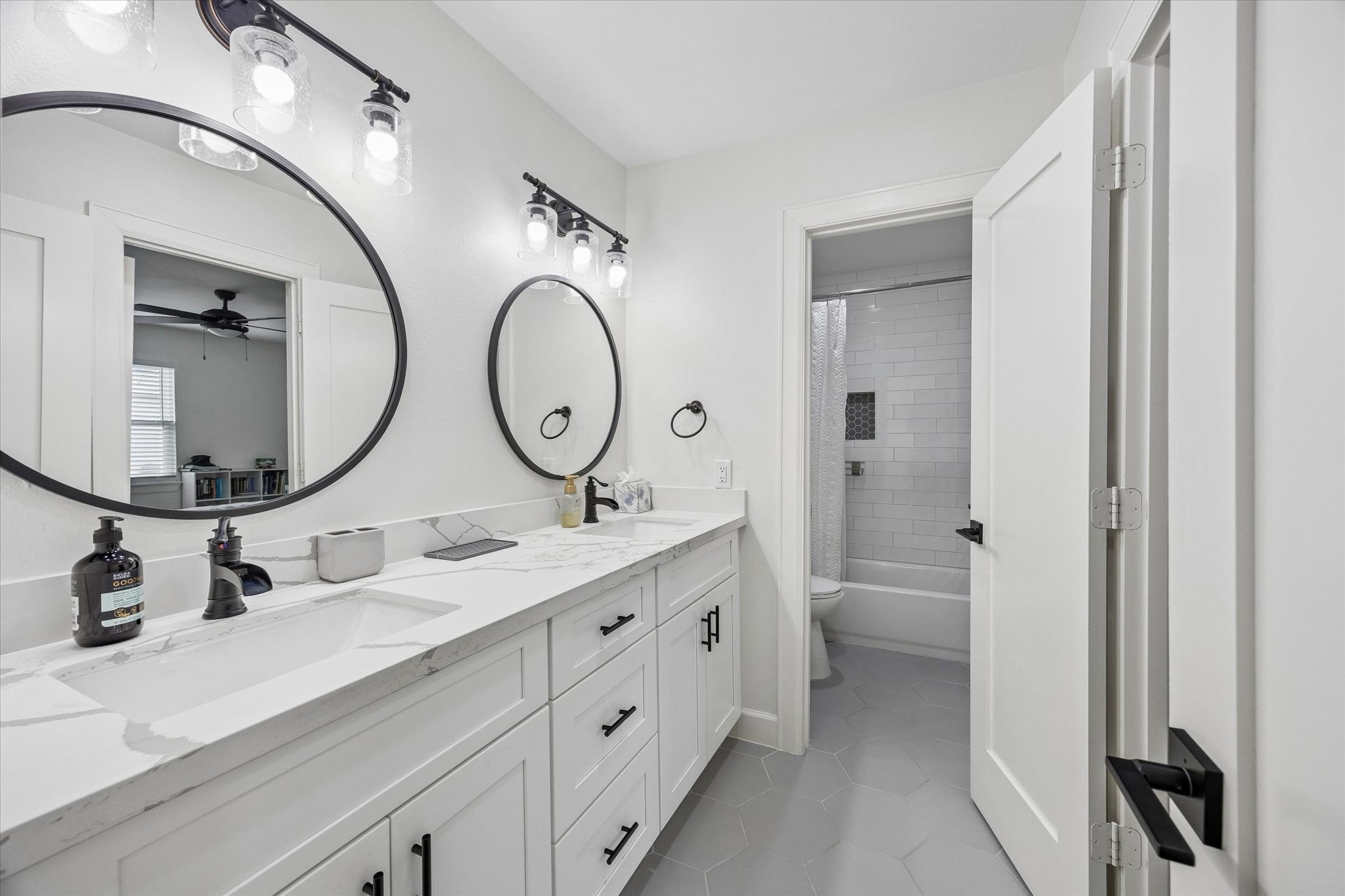 1420 Indiana Street Houston, TX 77006 - Photo 21 of 31 This bathroom features a modern double vanity with marble countertops, round mirrors, and stylish lighting fixtures. It has a clean, white aesthetic, hexagonal floor tiles, and includes a shower-tub combo with subway tile.
