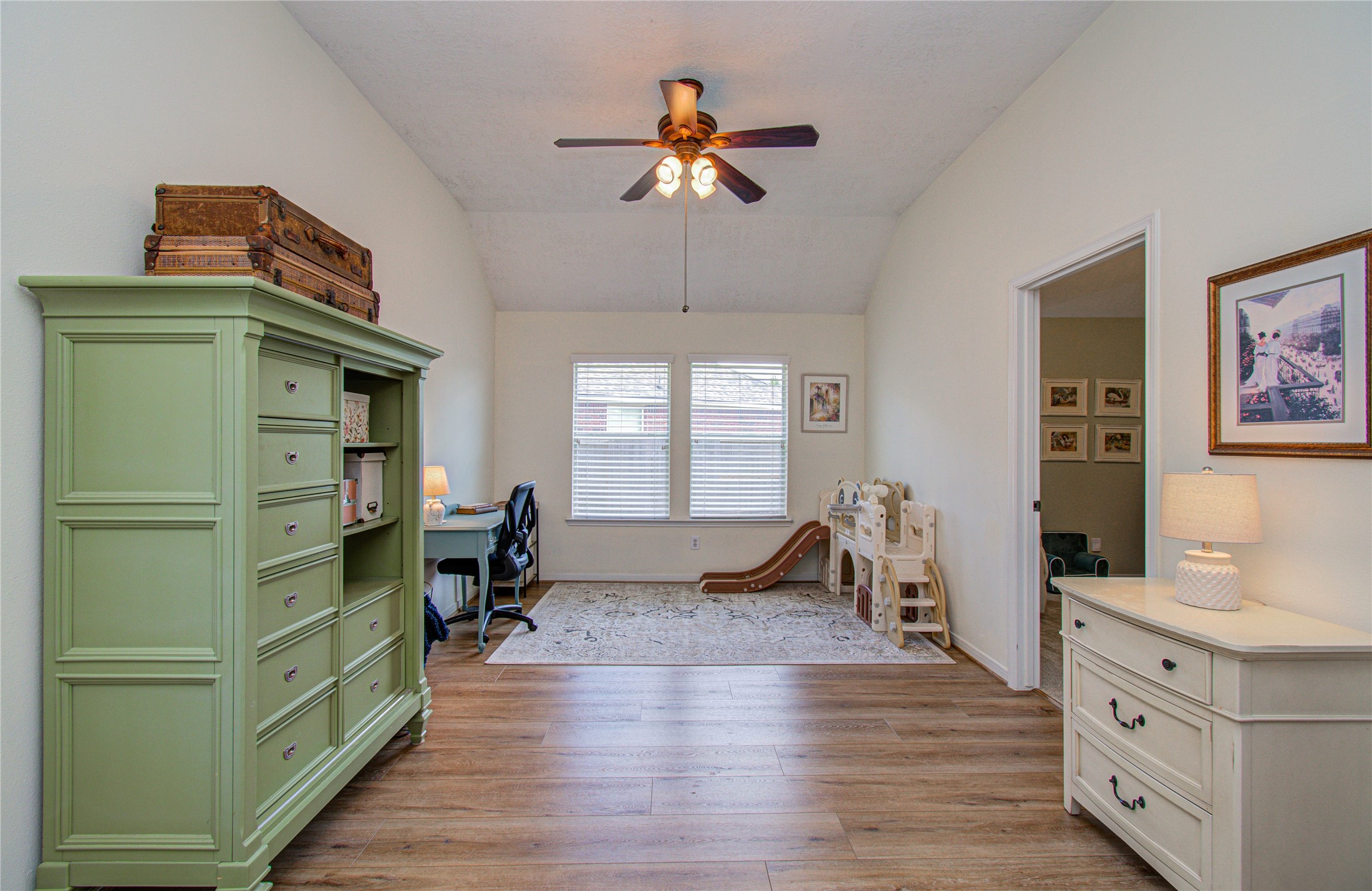 20007 Alcea Court Spring, TX 77379 - Photo 19 of 36 This room features a vaulted ceiling with a ceiling fan, wood flooring, and ample natural light from a central window. It includes a green dresser, a small desk, and a play area with a slide, making it versatile for both work and play.