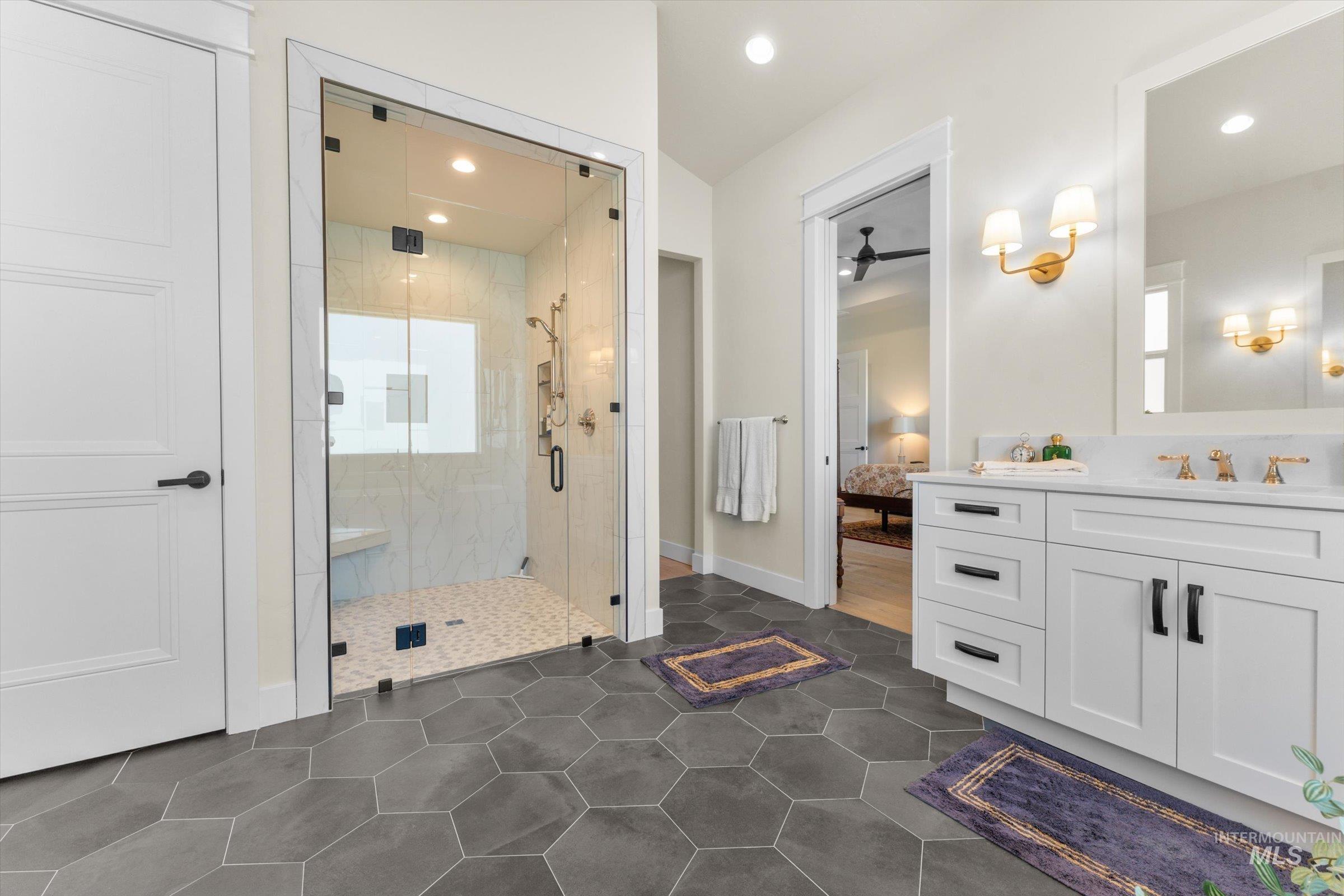 6585 South Pemberley Way Meridian, ID 83642 - Photo 26 of 43 Spacious bathroom featuring a glass-enclosed shower with white subway tile surround and bench, dark hexagon tile flooring, a white vanity with dual sinks and gold-finish fixtures, and a white interior door with dark hardware