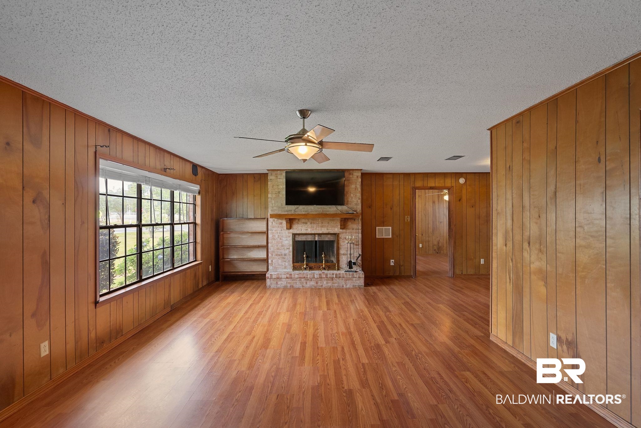 716 North Cedar Street Foley, AL 36535 - Photo 6 of 25 Spacious living area featuring wood-finish flooring, wood paneling, a brick fireplace with a mantel, built-in shelving, and a ceiling fan with integrated lighting