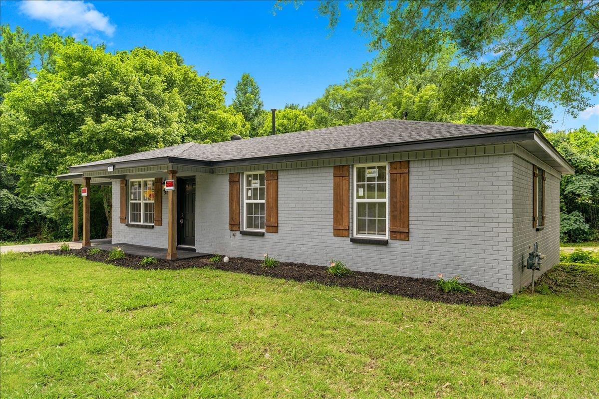 3065 Sax Road Memphis, TN 38109 - Photo 15 of 17 Gray brick exterior with dark wood shutters, a covered porch with wood columns, a dark shingle roof, and a landscaped front planter bed
