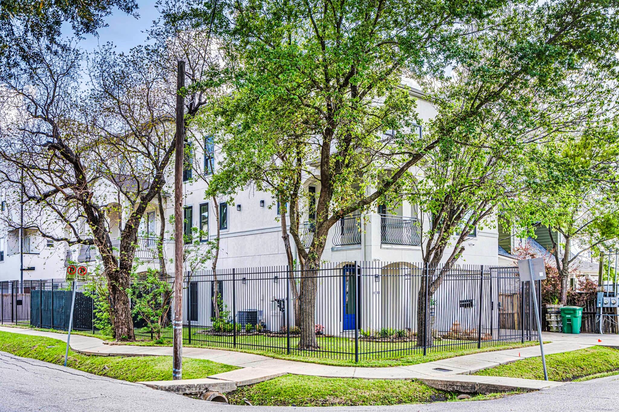 This property is a modern, multi-story home on a corner lot with a fenced yard and mature trees providing shade. The exterior features a clean, white facade with multiple balconies and large windows, offering a bright and airy feel. Located in a residential neighborhood, it combines privacy with easy access to surrounding areas.