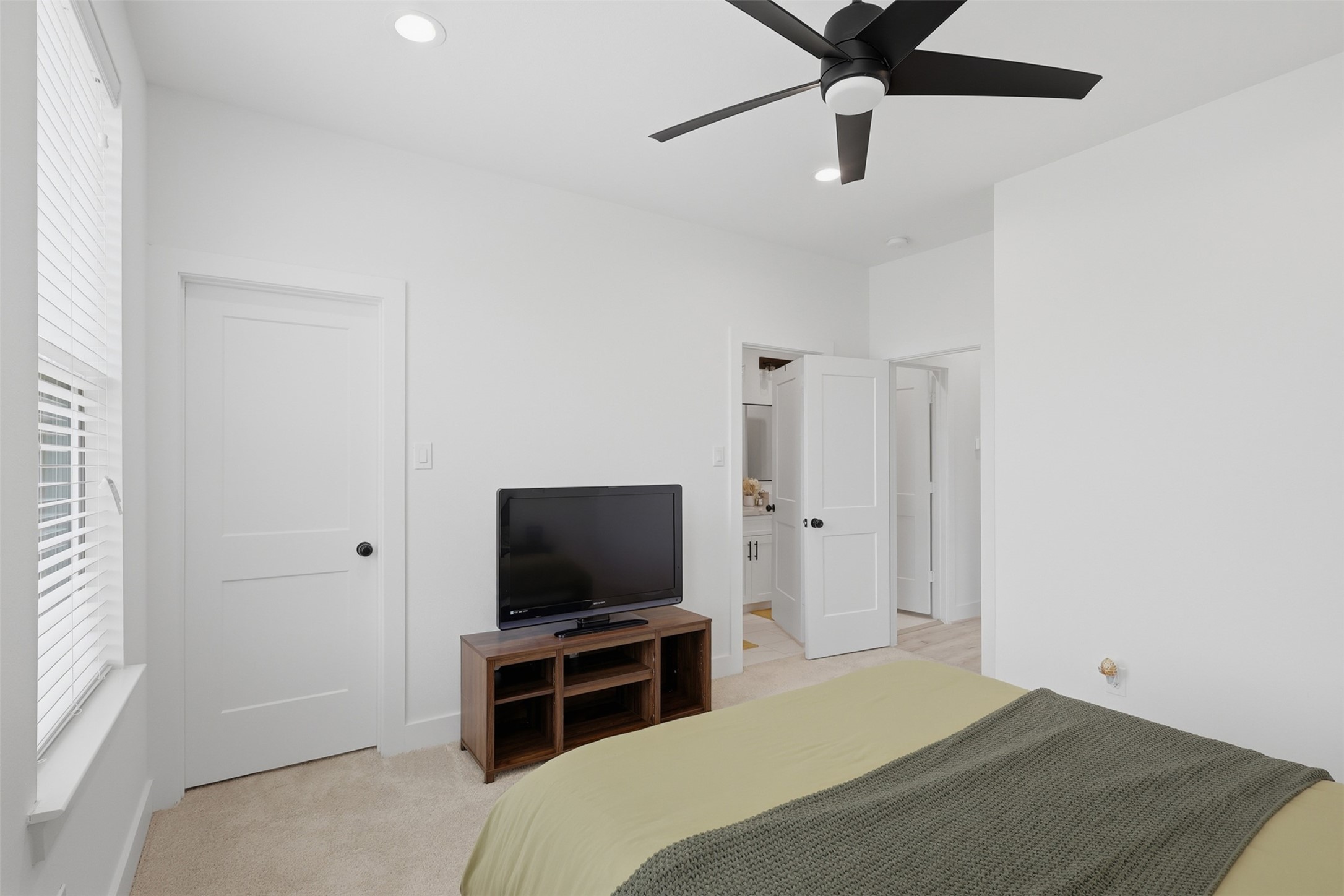 6833 Paris Street Houston, TX 77021 - Photo 16 of 23 This photo showcases a bright, modern bedroom with neutral walls, a ceiling fan, and ample natural light from a window. It features a cozy bed, a TV on a wooden stand, and an en-suite bathroom with white doors for a clean, inviting atmosphere.