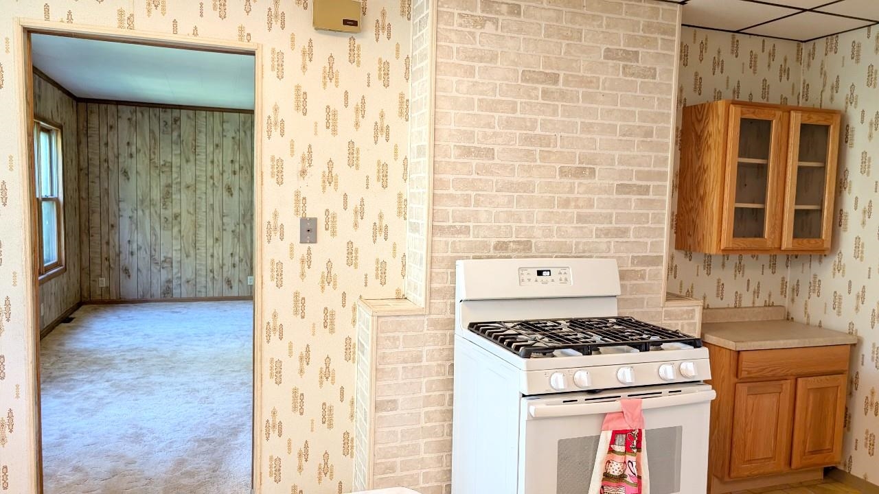 8159 Ludlow Road Cook, MN 55723 - Photo 15 of 32 Kitchen featuring white gas range oven, wallpapered walls, brown cabinets, light countertops, and glass insert cabinets