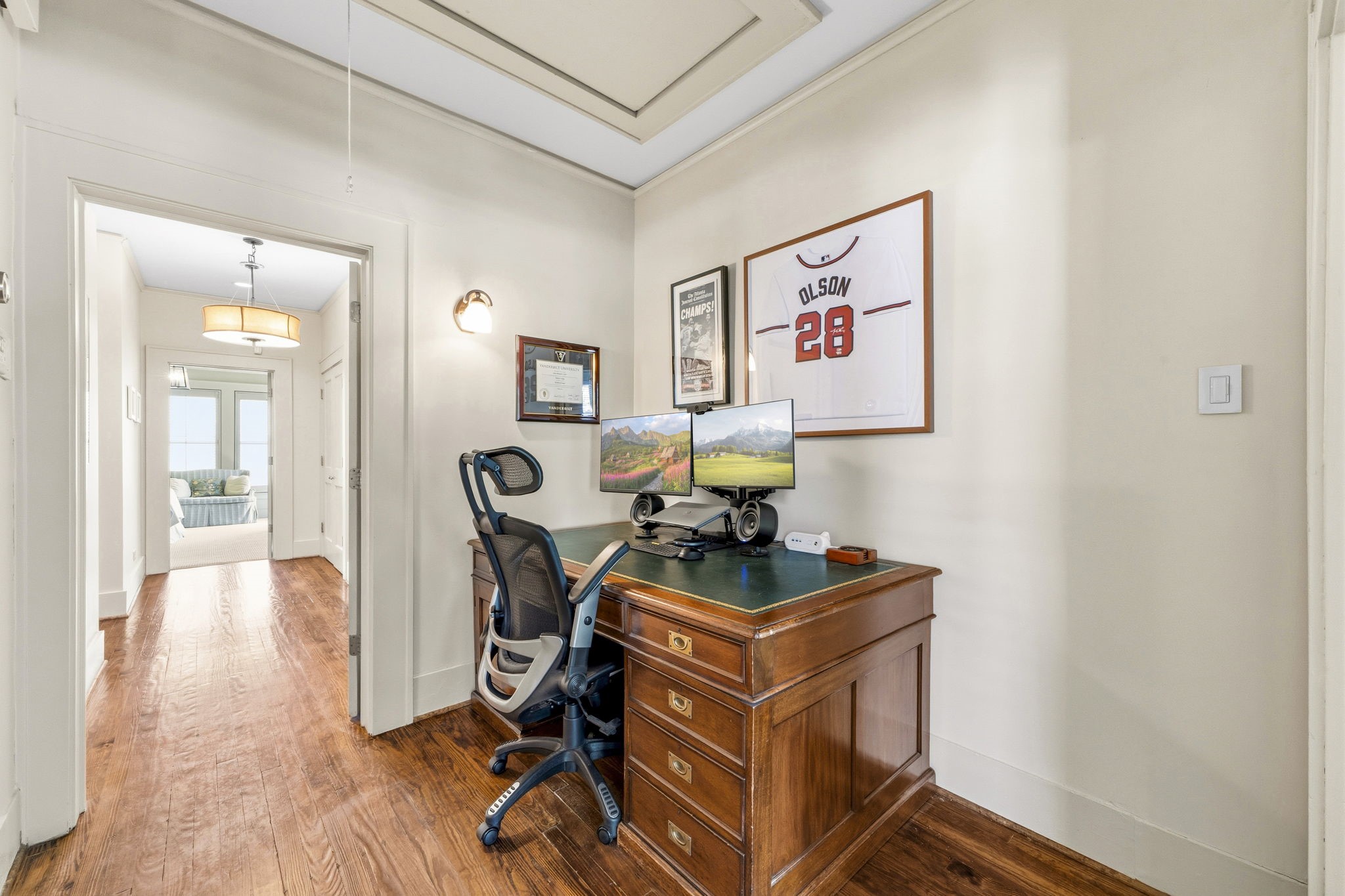 1521 Kipling Street Houston, TX 77006 - Photo 27 of 35 This photo showcases a cozy home office nook with a wooden desk and modern chair, set against a hallway with hardwood floors. The space is decorated with framed memorabilia and leads to a bright, inviting living area.