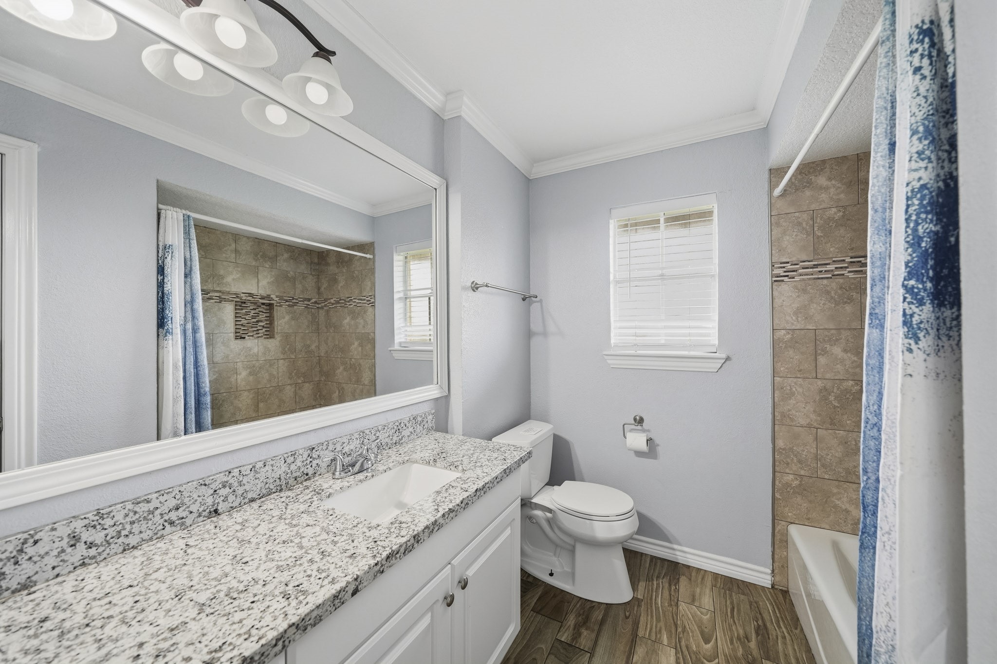 302 Country Road 893 Angleton, TX 77515 - Photo 18 of 26 This bathroom features a modern design with a granite countertop, white cabinetry, and wood-look tile flooring. It has a large mirror, updated light fixtures, and a shower-tub combo with stylish tile work. Natural light filters through a small window, enhancing the bright, fresh ambiance.