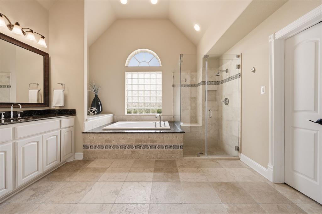 7013 Handel Colleyville, TX 76034 - Photo 20 of 37 Bathroom featuring a vaulted ceiling, a built-in tub with stone surround, a glass-enclosed shower, a double vanity with dark countertop, and large format tile flooring