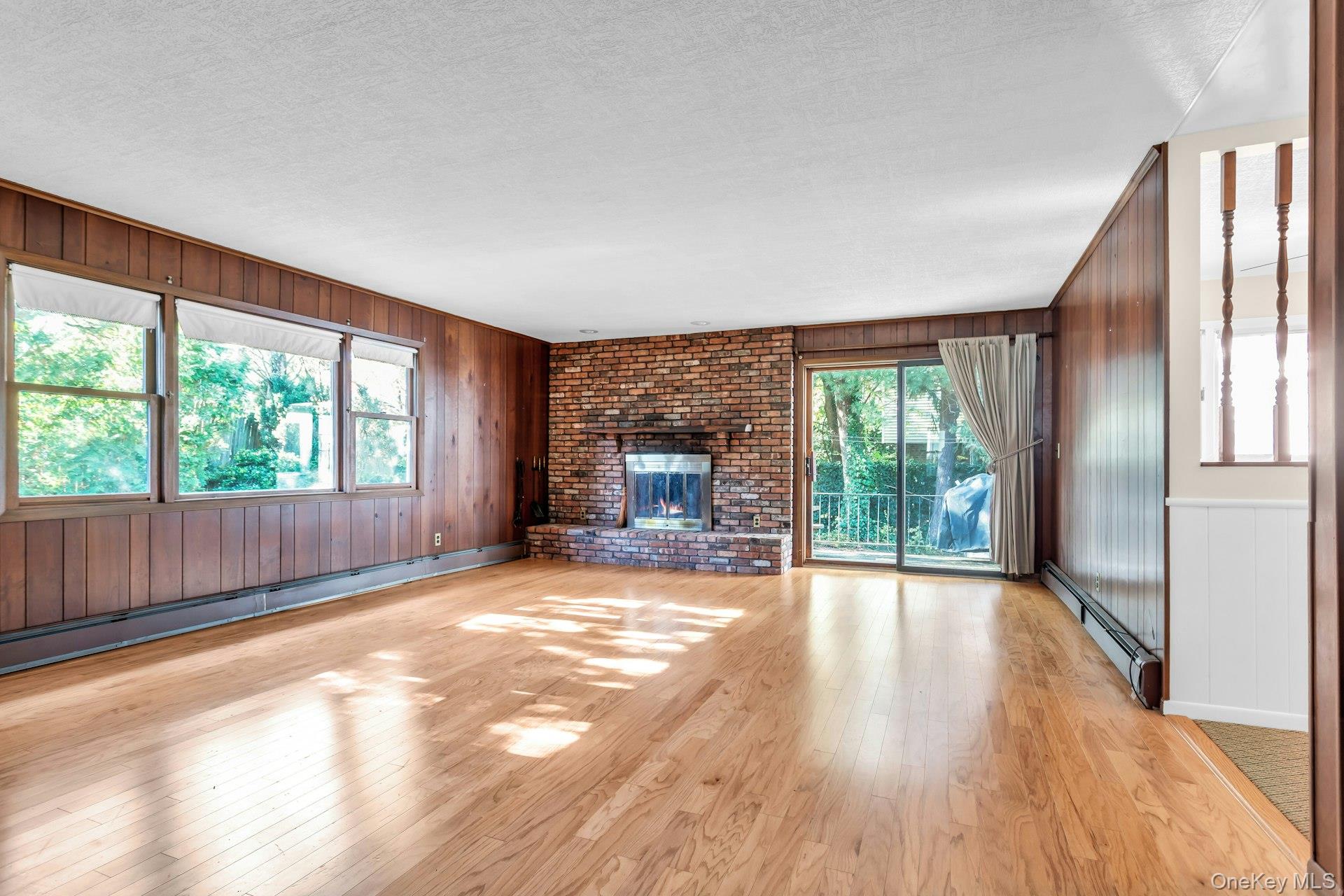 12 Violet Road Bayville, NY 11709 - Photo 14 of 42 Unfurnished living room featuring wooden walls, baseboard heating, a fireplace, light wood-type flooring, and a textured ceiling