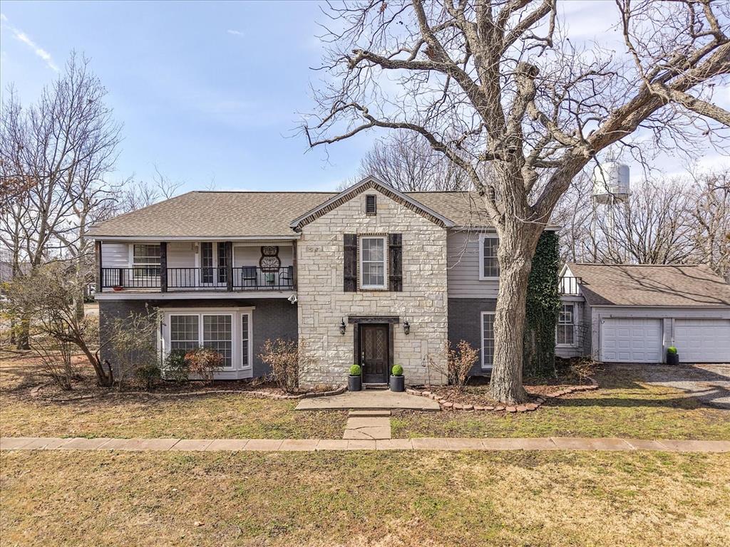 Traditional-style home with a shingled roof, a front lawn, stone siding, a balcony, and a garage