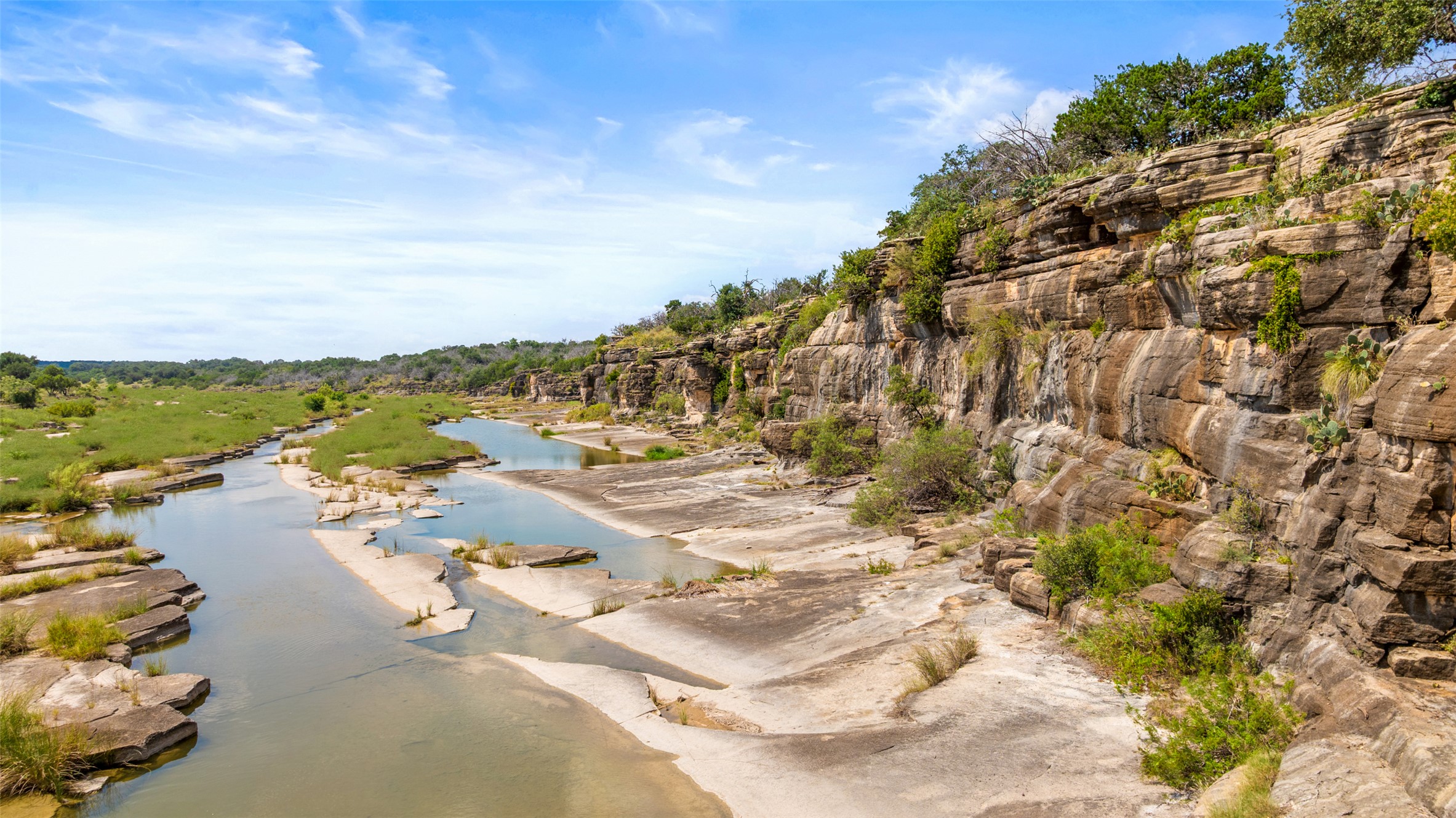 960 Gibson Road Johnson City, TX 78636 - Photo 2 of 19 A tranquil river winds through a rugged canyon, flanked by verdant banks and dramatic rock formations.