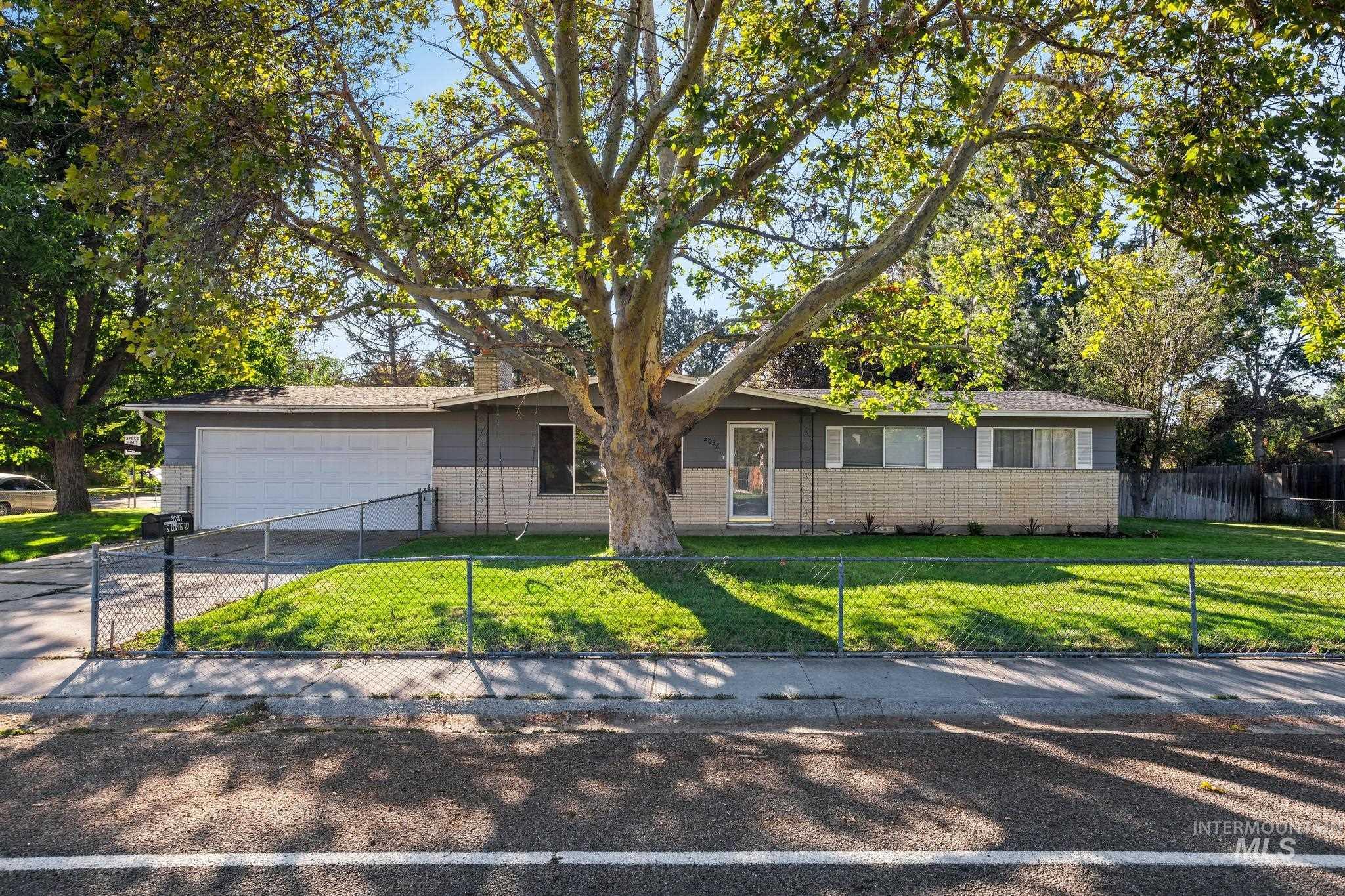 Single-story residence featuring a two-car garage, front yard with mature trees, and a chain-link fence