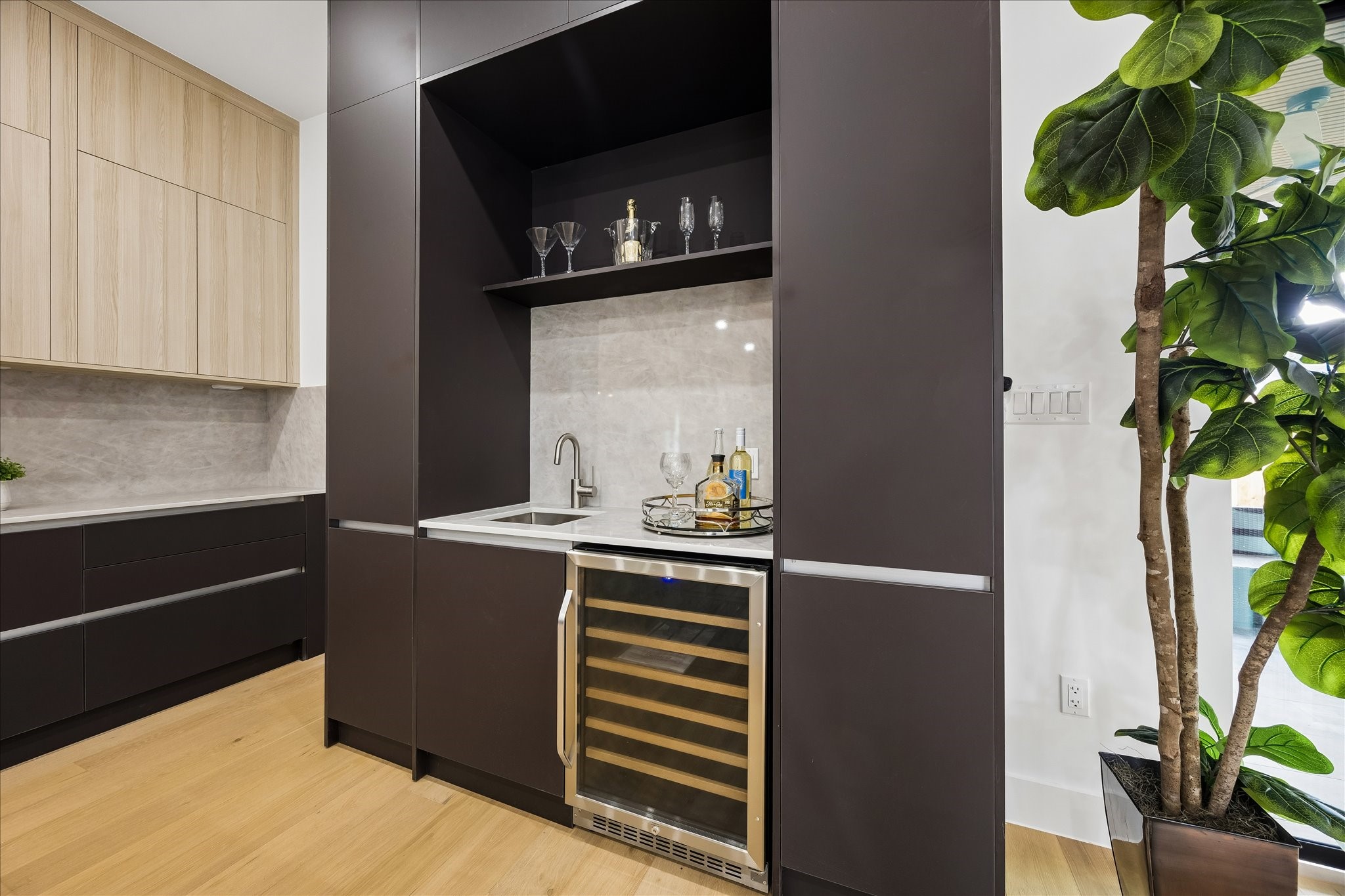 5430 Rutherglenn Drive Houston, TX 77096 - Photo 10 of 31 Private wine bar area with sleek dark cabinetry, a built-in wine cooler, and a compact sink. The contrasting wood cabinets and light hardwood flooring add warmth, while the marble backsplash and open shelving provide a touch of elegance.