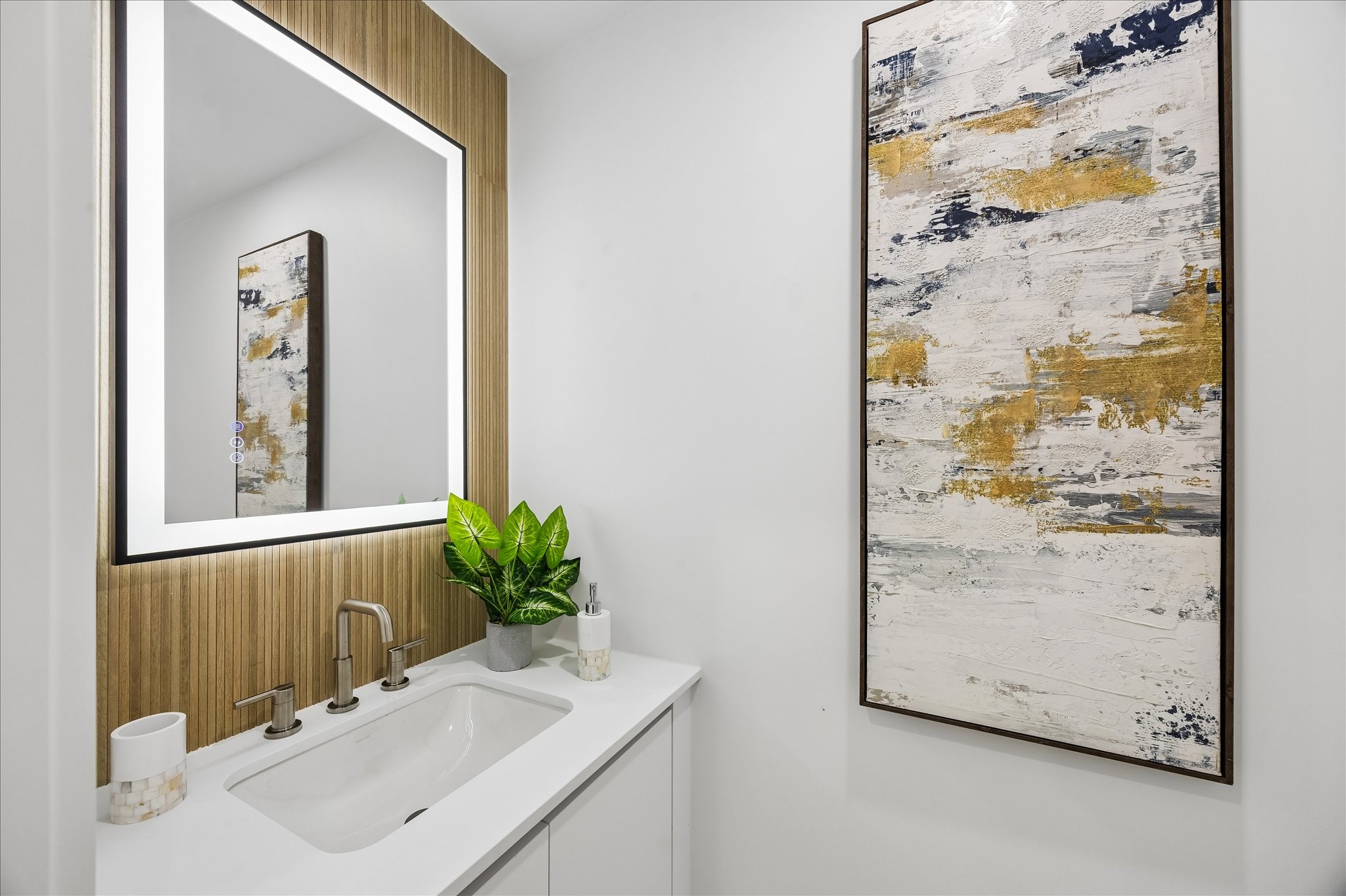 5430 Rutherglenn Drive Houston, TX 77096 - Photo 12 of 31 The downstairs half bathroom features a modern design with a sleek white vanity, under-mounted sink, and contemporary faucet. The large, backlit mirror enhances the space, complemented by a textured wooden accent wall.