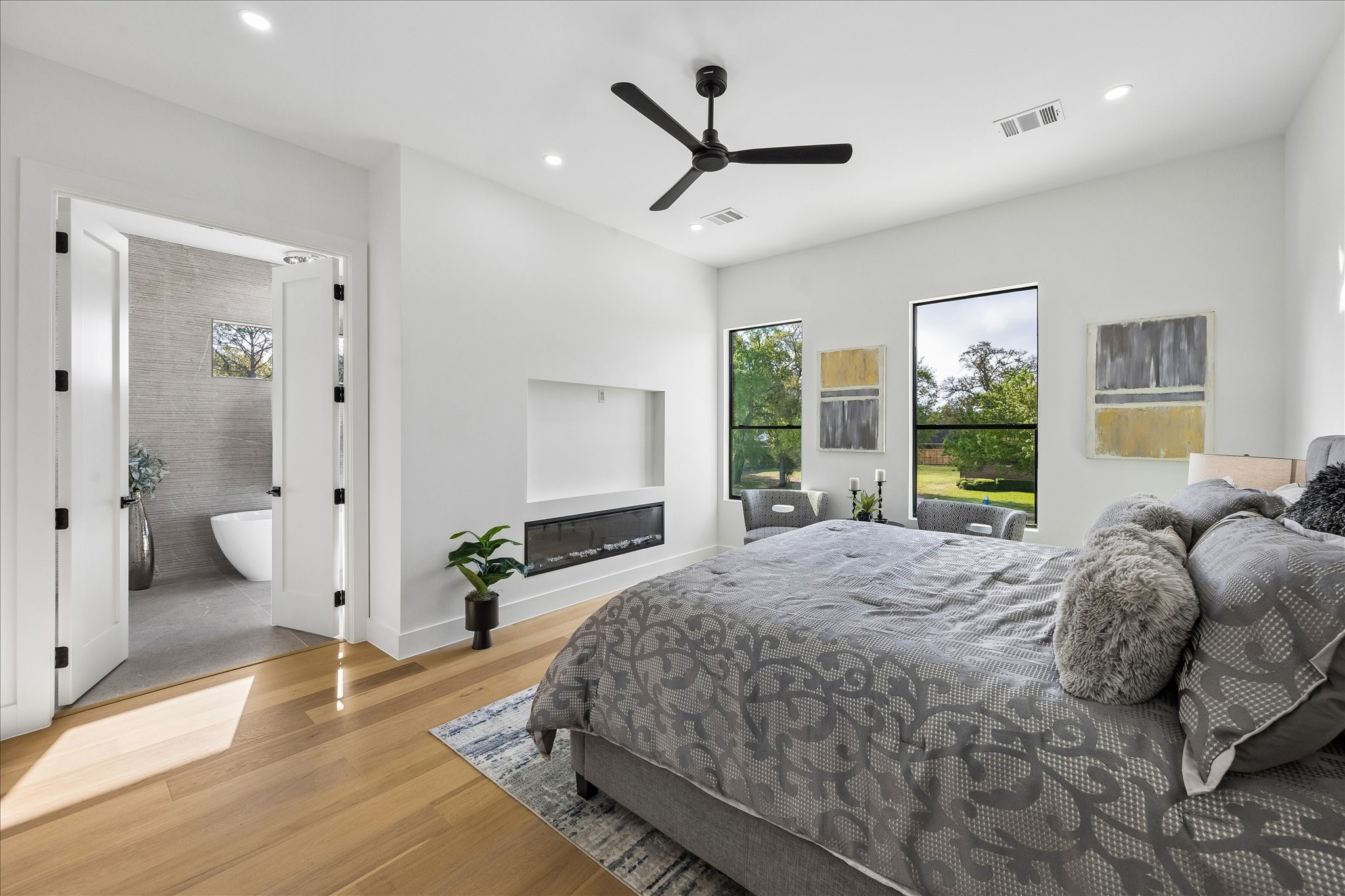 5430 Rutherglenn Drive Houston, TX 77096 - Photo 15 of 31 The primary bedroom features large windows with garden views, and a sleek built-in fireplace. The room connects to a stylish en-suite bathroom with a freestanding tub, all accented by elegant decor and light wood flooring.