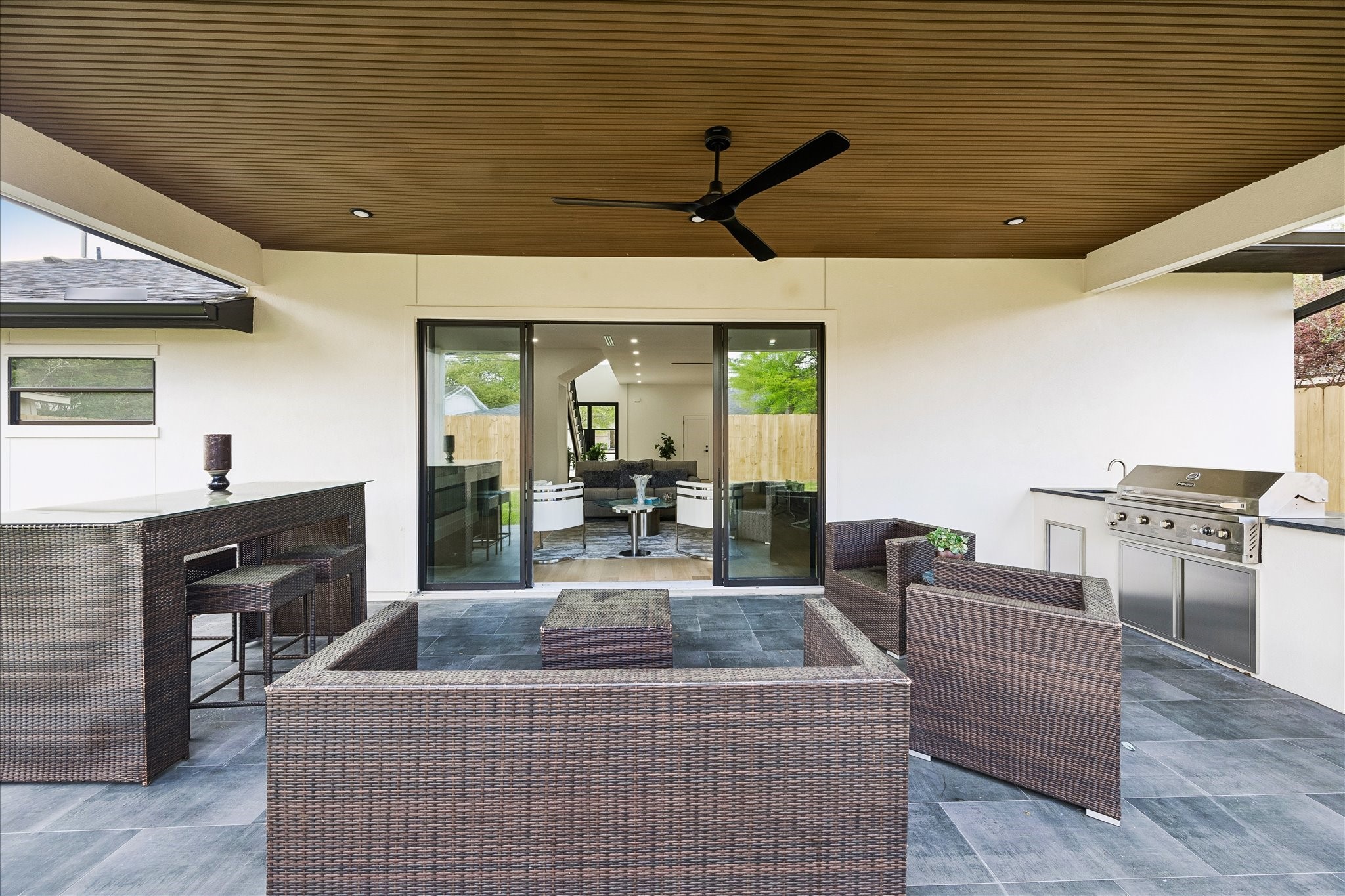 5430 Rutherglenn Drive Houston, TX 77096 - Photo 24 of 31 This outdoor patio features a sleek, modern design with a cozy wicker seating area, a built-in grill, and a bar counter. The space is covered with a wood-paneled ceiling and a ceiling fan, offering a seamless indoor-outdoor transition through large sliding glass doors. Perfect for entertaining.