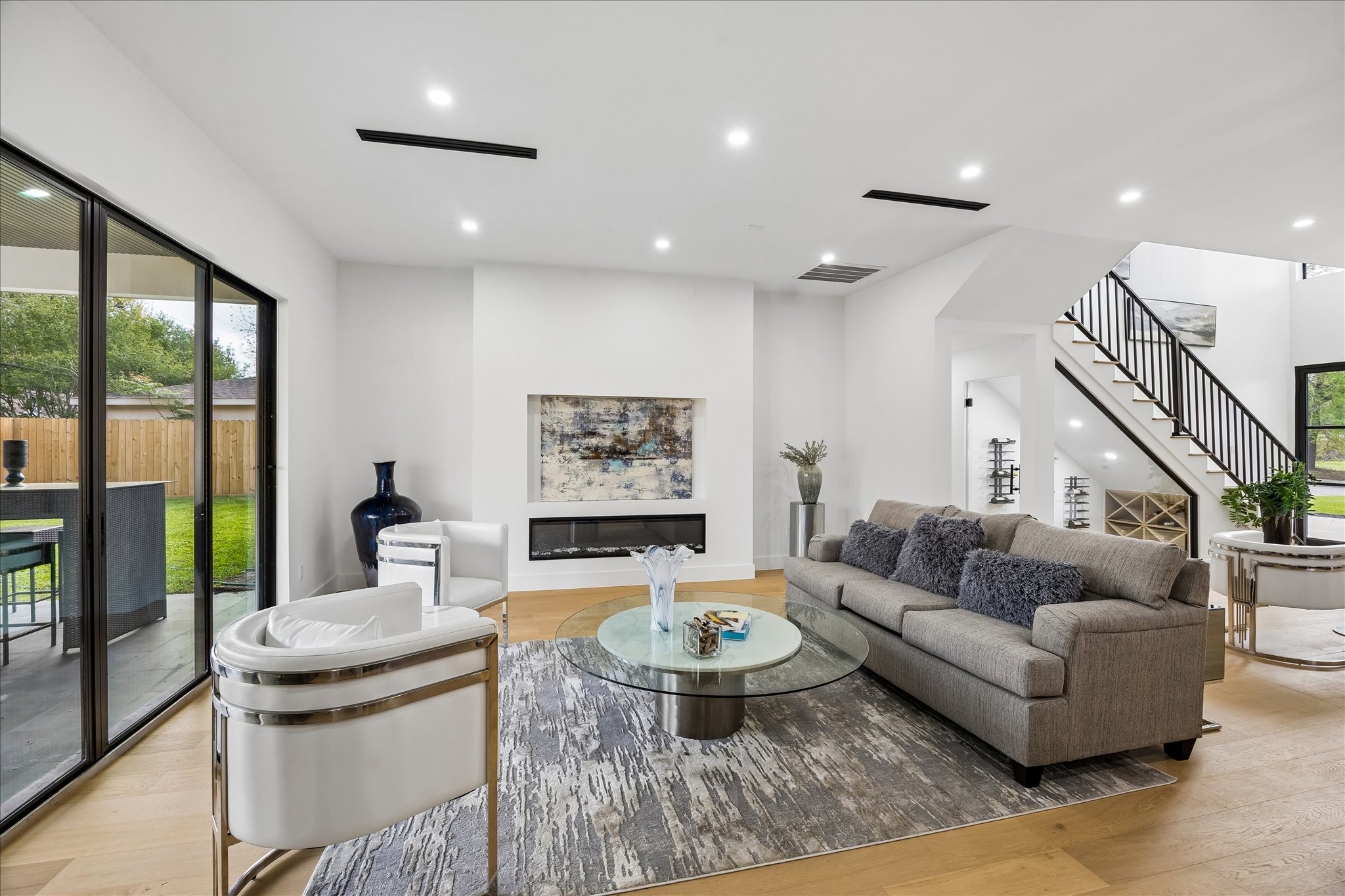 5430 Rutherglenn Drive Houston, TX 77096 - Photo 7 of 31 This modern living room features a spacious open-plan design with large glass doors leading to a patio, allowing ample natural light. With sleek furnishings, a cozy fireplace, and a stylish staircase, create a welcoming and contemporary atmosphere to enjoy.