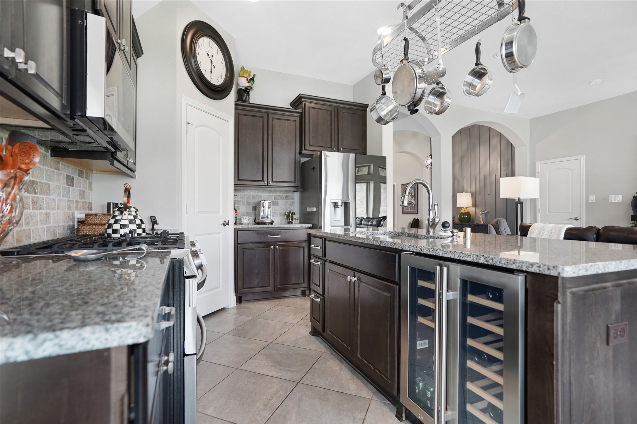 2345 Redwood Ridge Trail Manvel, TX 77578 - Photo 16 of 39 This modern kitchen features dark wood cabinetry, granite countertops, and stainless steel appliances. A central island with a built-in wine fridge and a hanging pot rack adds functionality, while the open layout flows into a spacious living area, enhancing the home's inviting atmosphere.