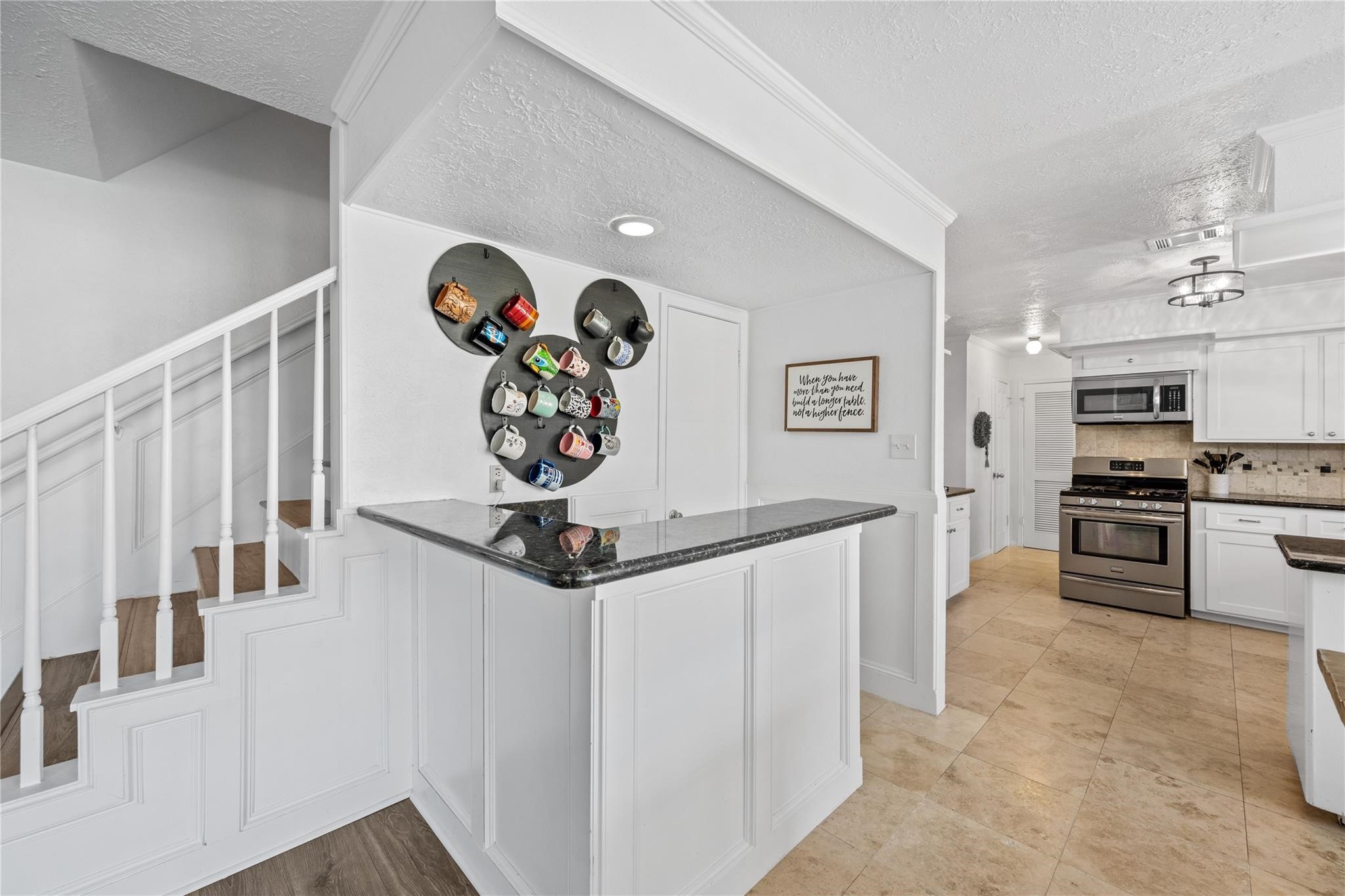 19702 Sweet Forest Lane Humble, TX 77346 - Photo 13 of 30 This photo showcases a modern kitchen with stainless steel appliances, white cabinetry, and tiled flooring. A unique mug display adds character, while an adjacent staircase leads to the upper level. The space is bright and inviting, ideal for both cooking and entertaining.