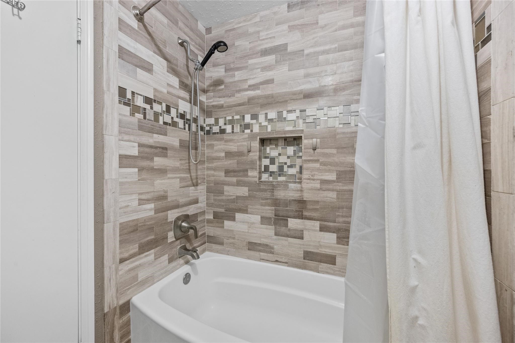 19702 Sweet Forest Lane Humble, TX 77346 - Photo 23 of 30 A modern bathroom features a tiled shower and bathtub combination with earth-toned, textured tiles and a decorative mosaic accent. A built-in niche adds convenience and style.