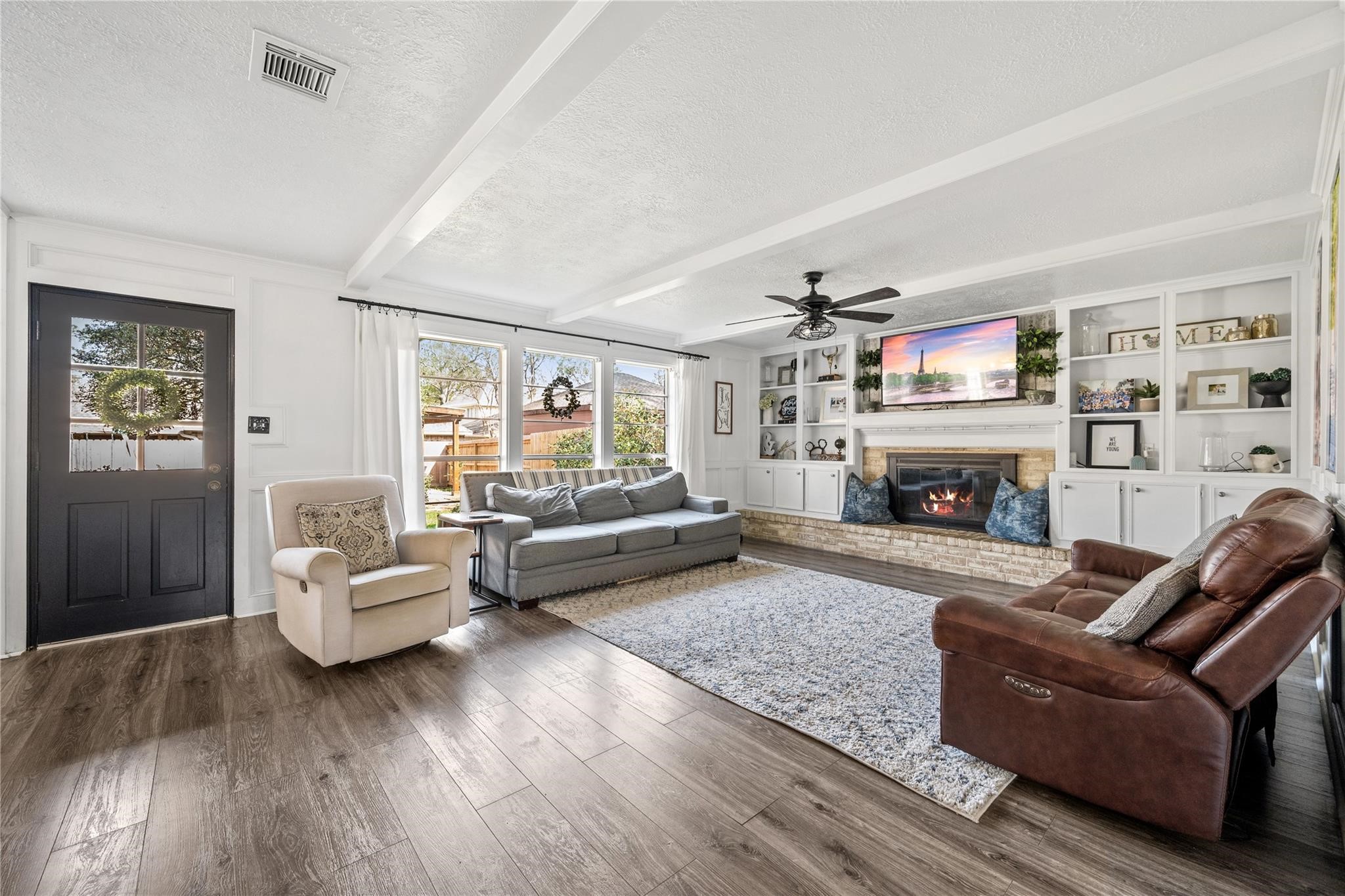 19702 Sweet Forest Lane Humble, TX 77346 - Photo 7 of 30 This inviting living room features a cozy fireplace, built-in shelving, and abundant natural light from large windows. The space is enhanced by a ceiling fan, wood flooring, and a comfortable seating arrangement, creating a welcoming atmosphere.