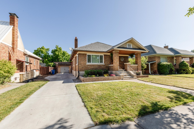 227 West 200 North Logan, UT 84321 - Photo 3 of 44 Brick exterior with a covered front porch featuring stone steps and wrought iron railings, a concrete driveway leading to a detached garage, and a manicured front lawn with established landscaping