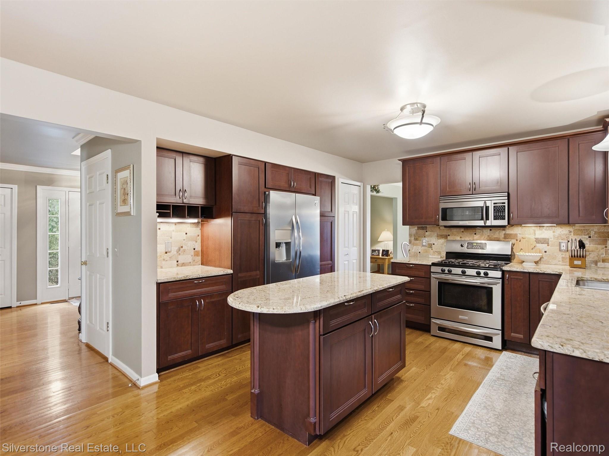 6577 Edith Court Troy, MI 48085 - Photo 21 of 61 Kitchen featuring an island with a curved countertop, wood-finish flooring, stainless steel appliances, dark wood cabinetry, and a stone tile backsplash