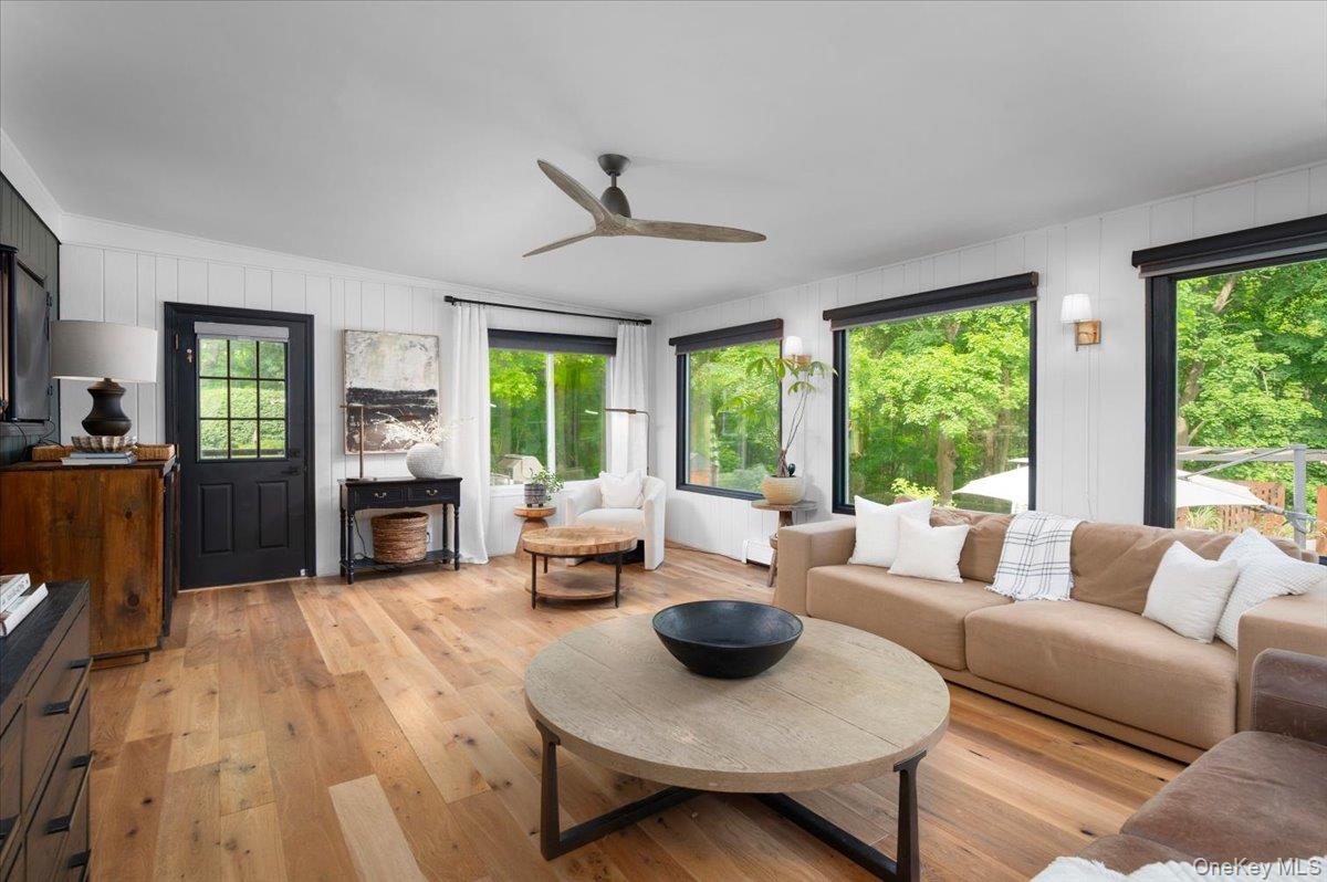 13 Jacks Pond Road Newburgh, NY 12550 - Photo 16 of 44 Airy living space showcasing hardwood flooring throughout, white shiplap walls, a ceiling fan, a black grid-paneled door, and several large black-framed windows.