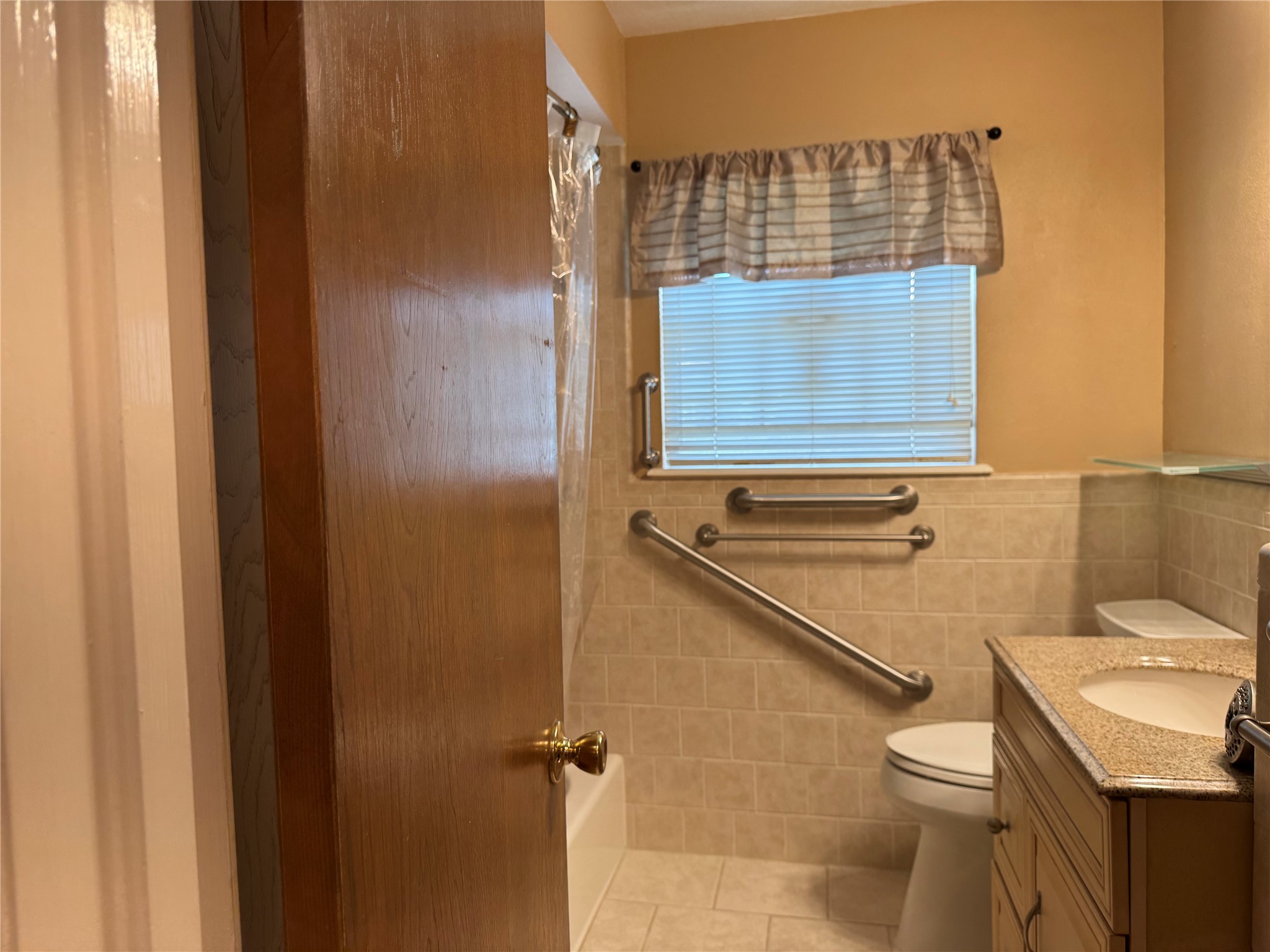 203 East Thornton Road Houston, TX 77022 - Photo 10 of 18 This bathroom features a tiled shower with a grab bar, a window with a valance, and a wooden vanity with a granite countertop. The space is accessible and has a cozy, functional design.